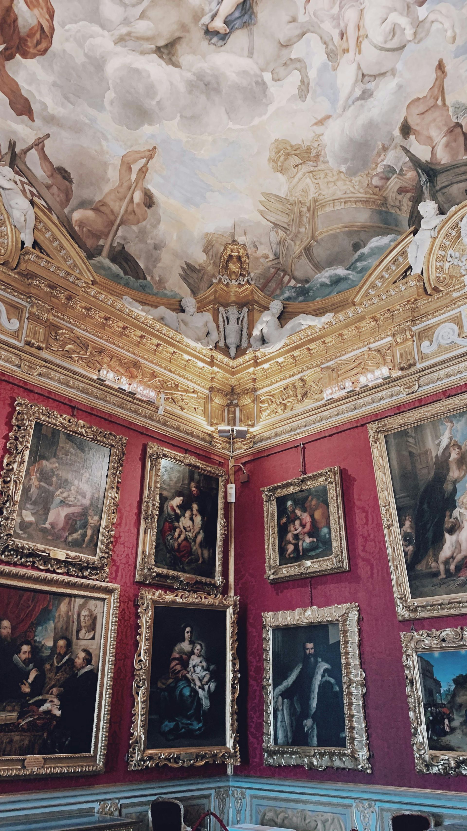 Ornate room with red walls, gold trim, and ceiling fresco. Paintings line the walls, a classical setting.
