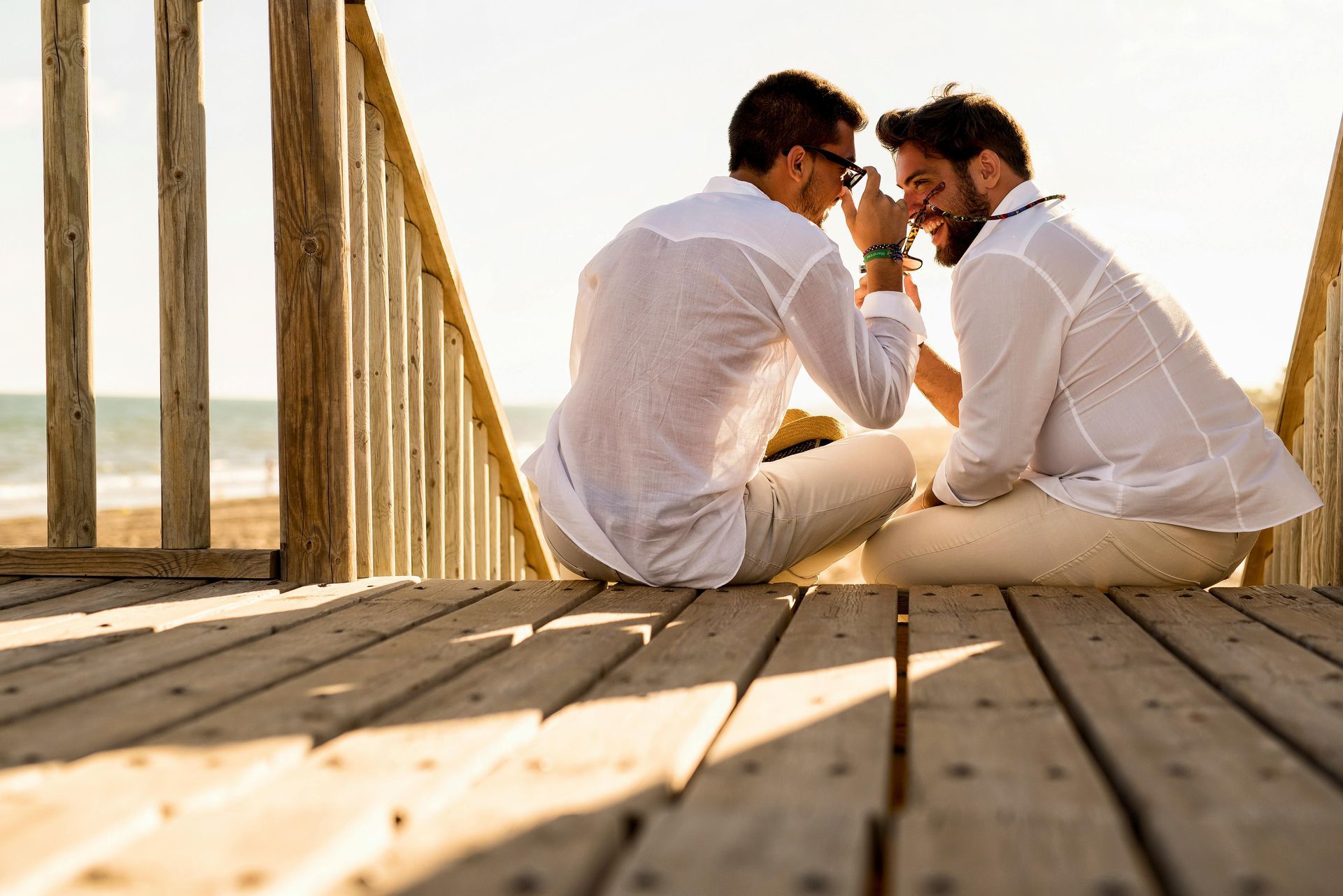 Two men in white shirts and pants sit close together on a wooden deck, facing each other, possibly at the beach. Gay Holidays to Spain