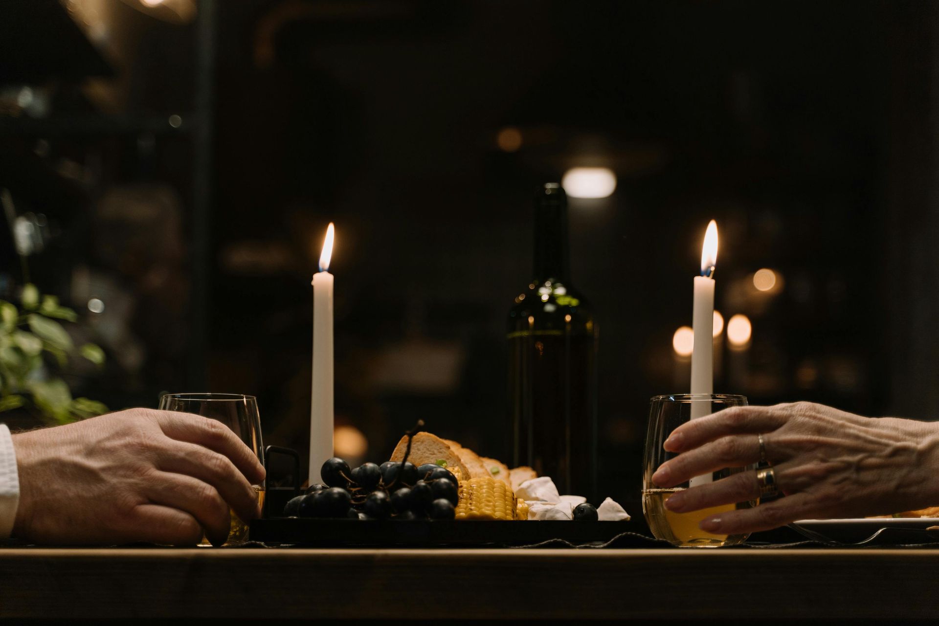 Two people at candlelit dinner, holding drinks. Food, wine bottle on table. Dark setting.