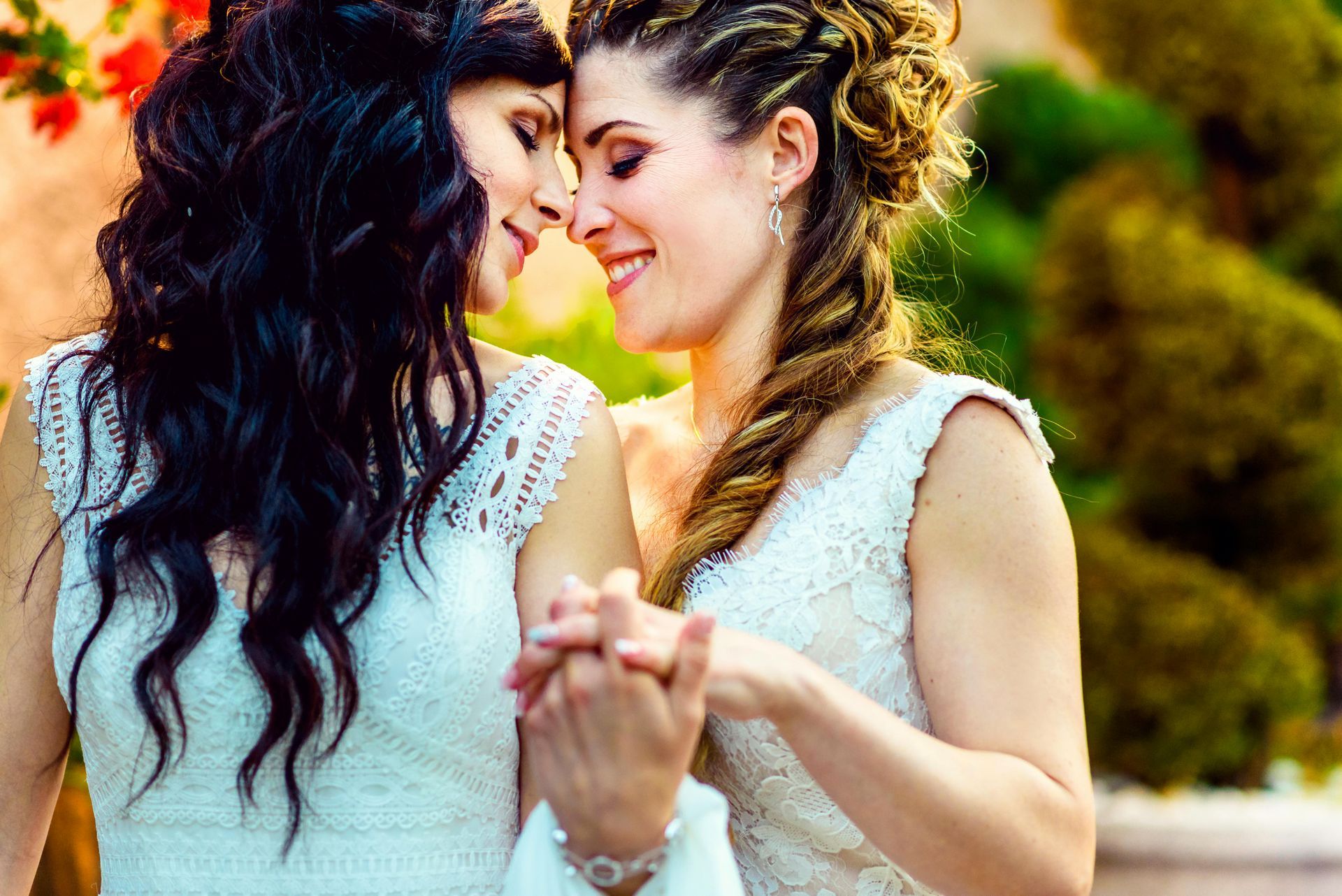 Two women in wedding dresses, smiling and holding hands, foreheads touching in an outdoor setting.