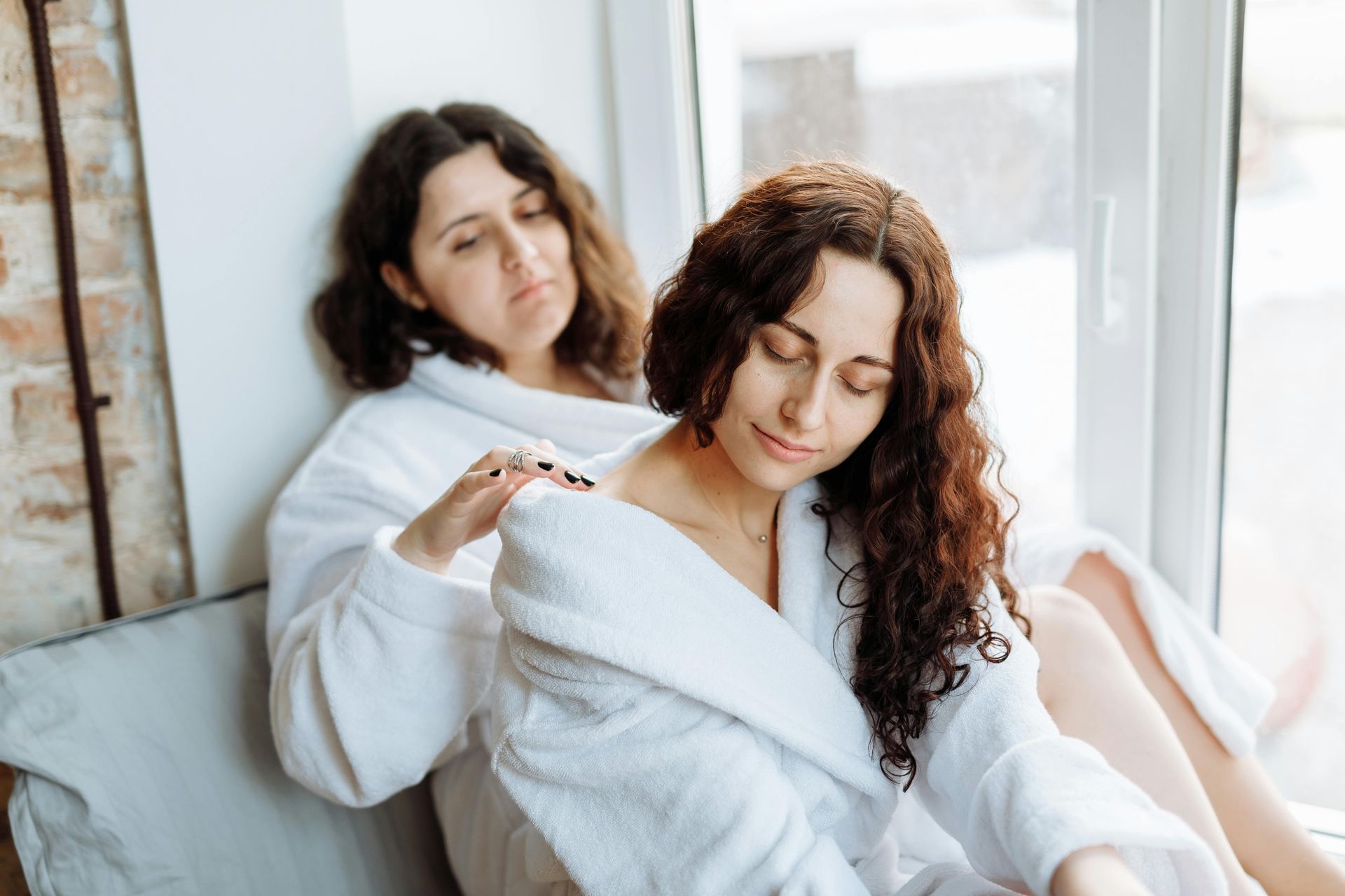 Two women in white robes: one massages the other's shoulders near a window.