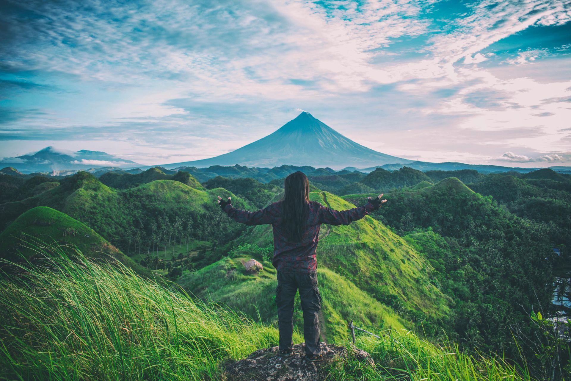 Person with arms outstretched atop green hills, mountain in background, cloudy sky.