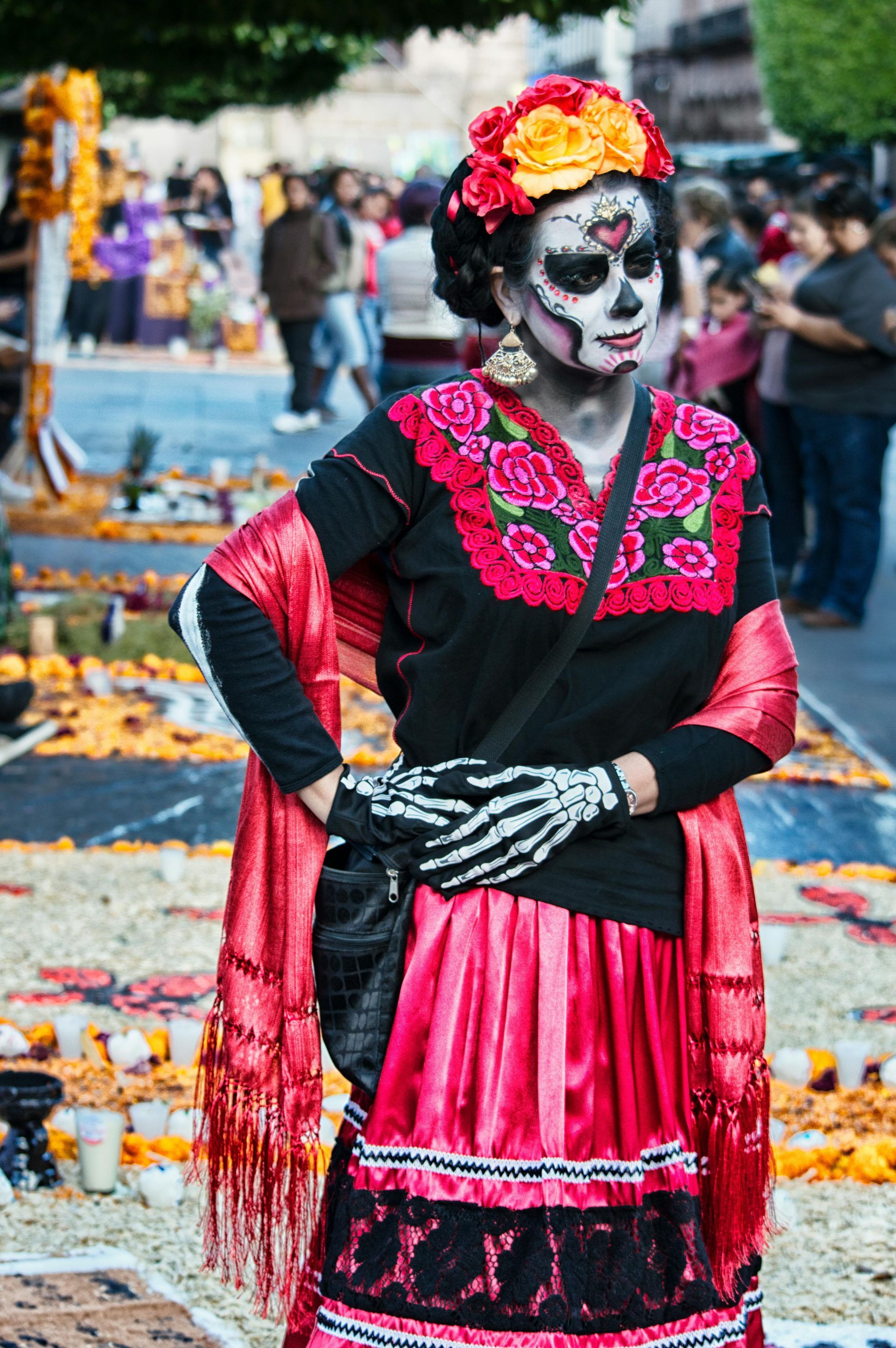 Woman in Día de los Muertos attire, black and pink embroidered dress, skeleton makeup, flowers in hair, standing outdoors.