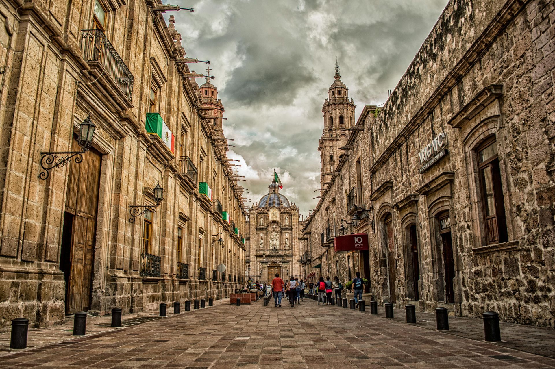 Stone buildings line a cobblestone street leading to a cathedral under a cloudy sky. People walk.