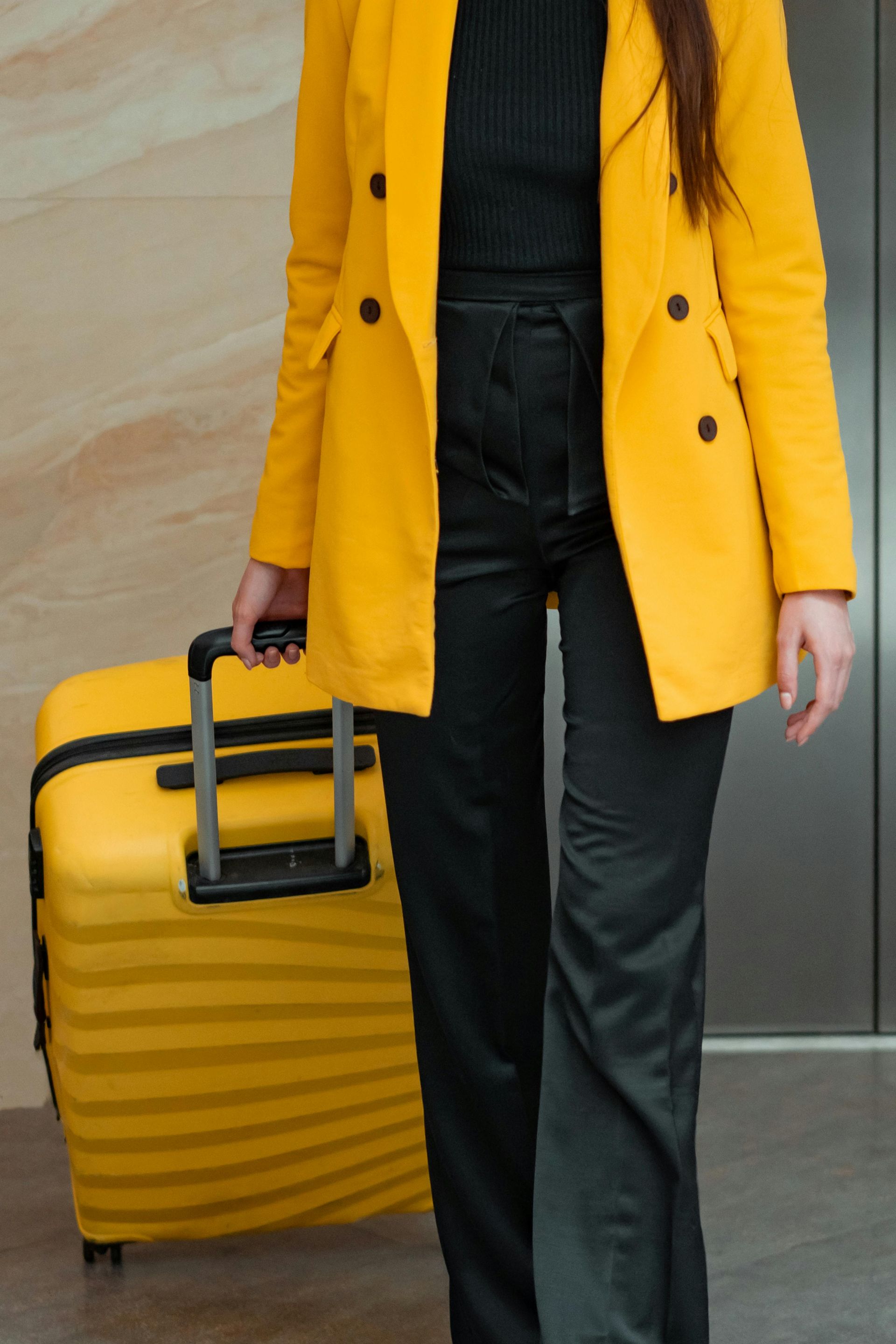 Woman in yellow blazer and black pants pulls a yellow suitcase. Elevator in background.