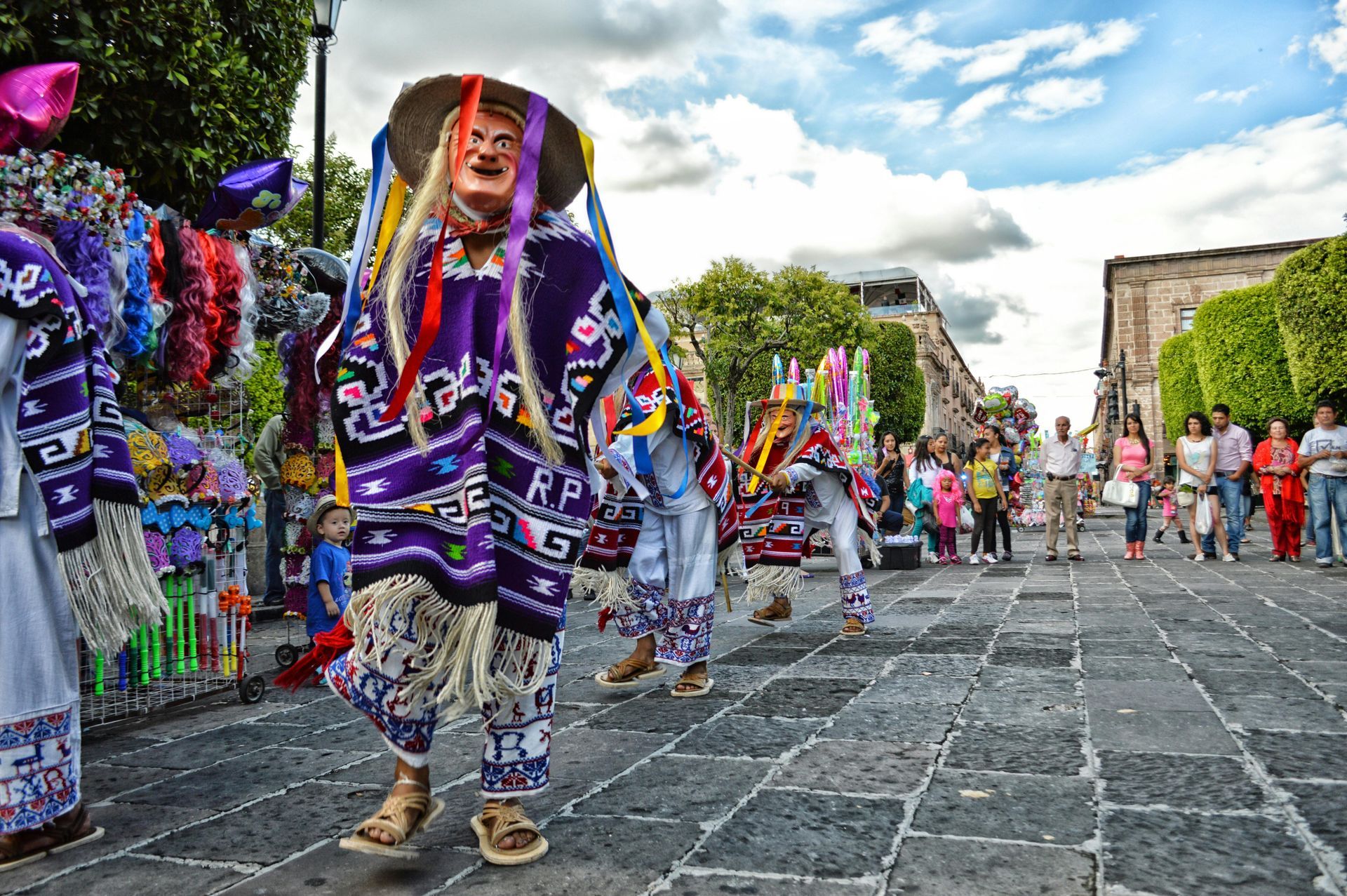 People in colorful costumes with masks dance on a cobblestone street, spectators watch in the background. Mexico