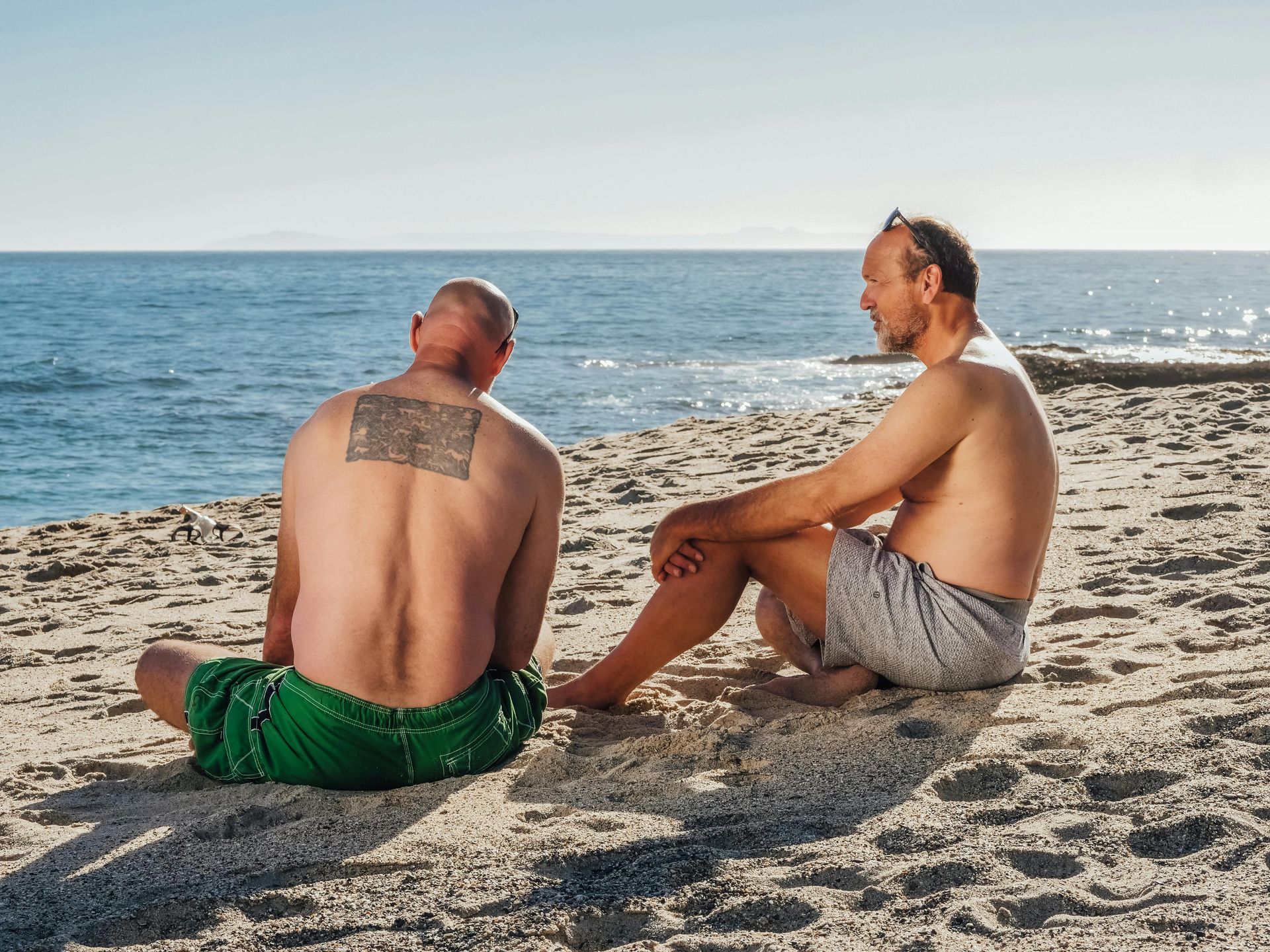 Two people sit on a sandy beach, facing each other with the ocean in the background.