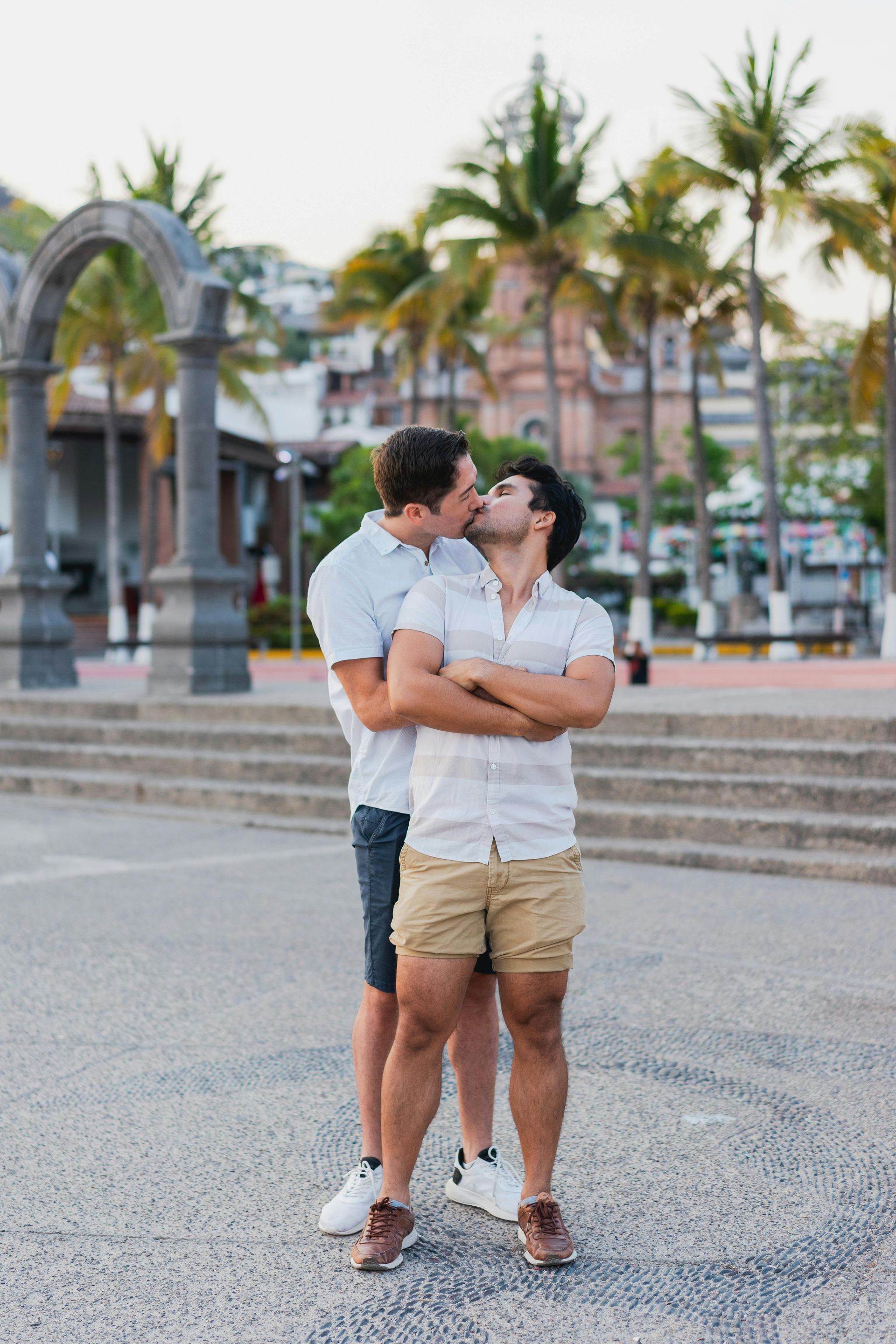 Two men kissing outdoors, arms crossed, by stone architecture and palm trees.