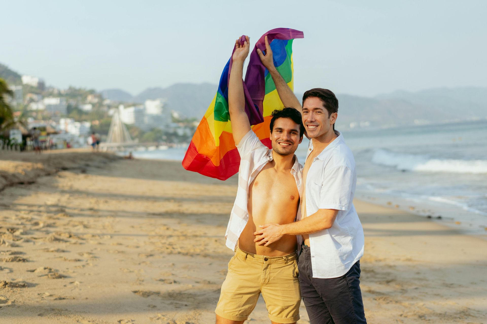 Two men on a beach, one holding a rainbow flag, the other hugs him, smiles, seaside view.