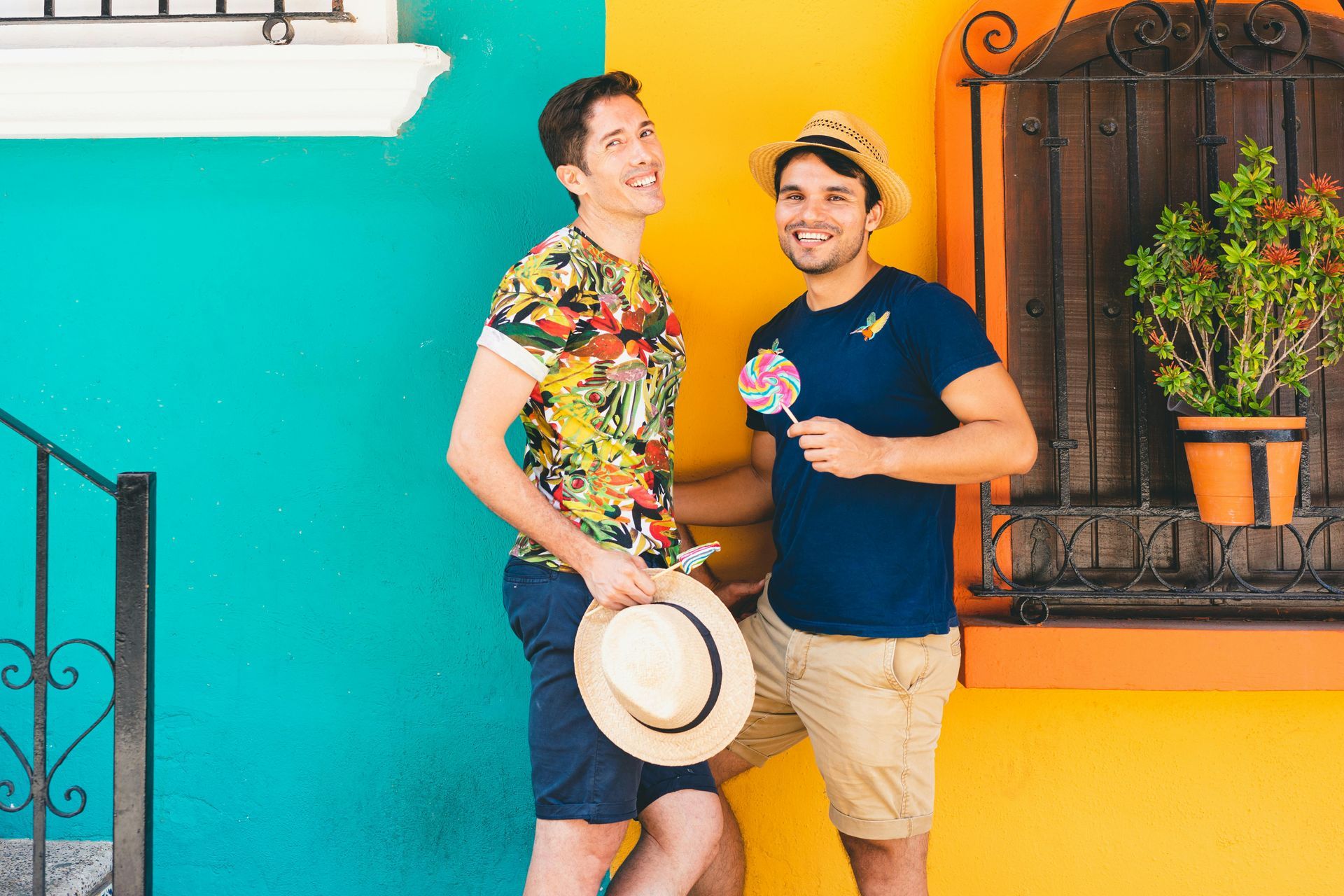 Two men pose smiling in front of colorful walls. One holds a hat, the other a lollipop.