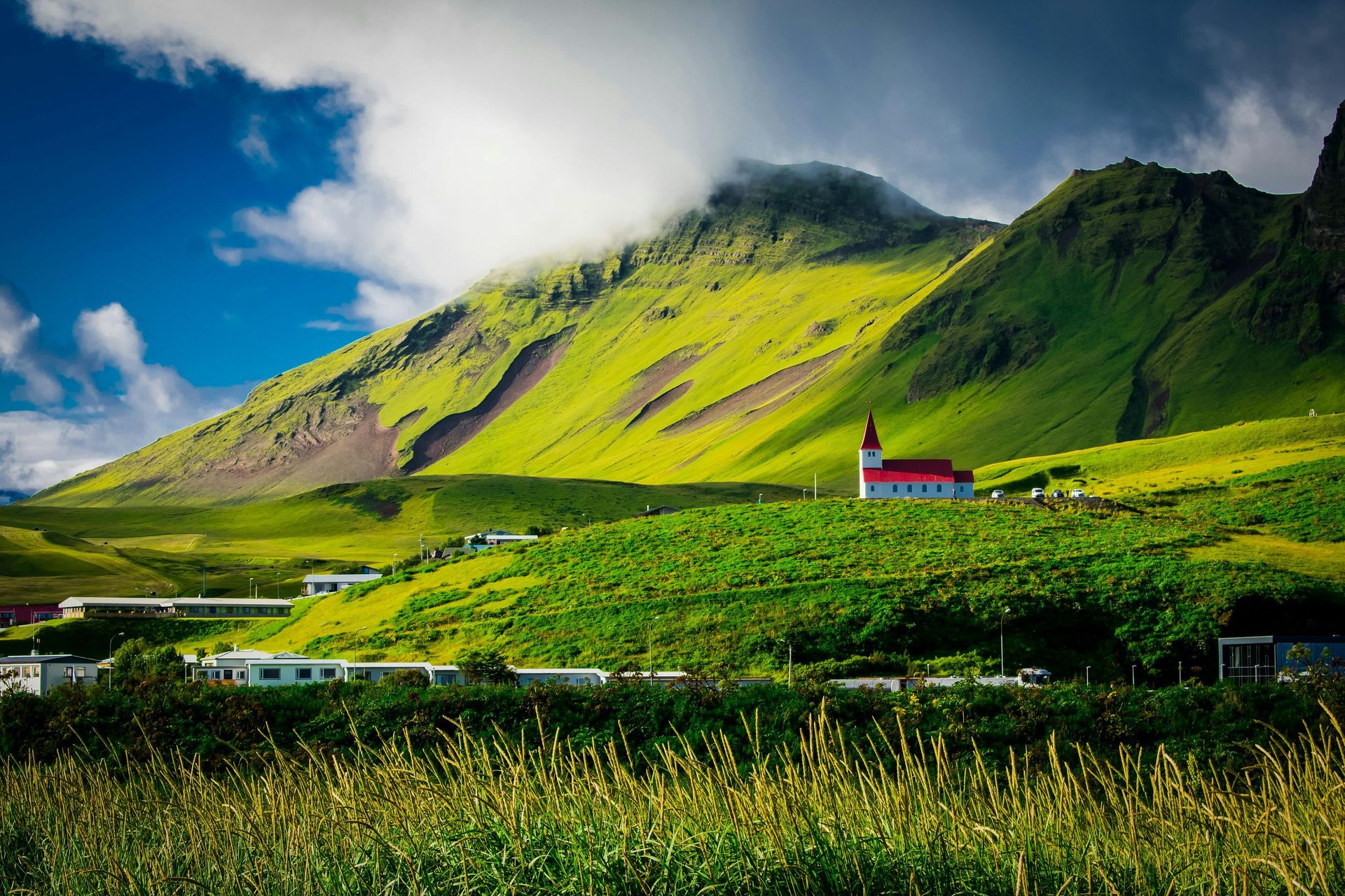 Green hills surround a white church with a red roof, set in a lush landscape under a blue sky.Iceland