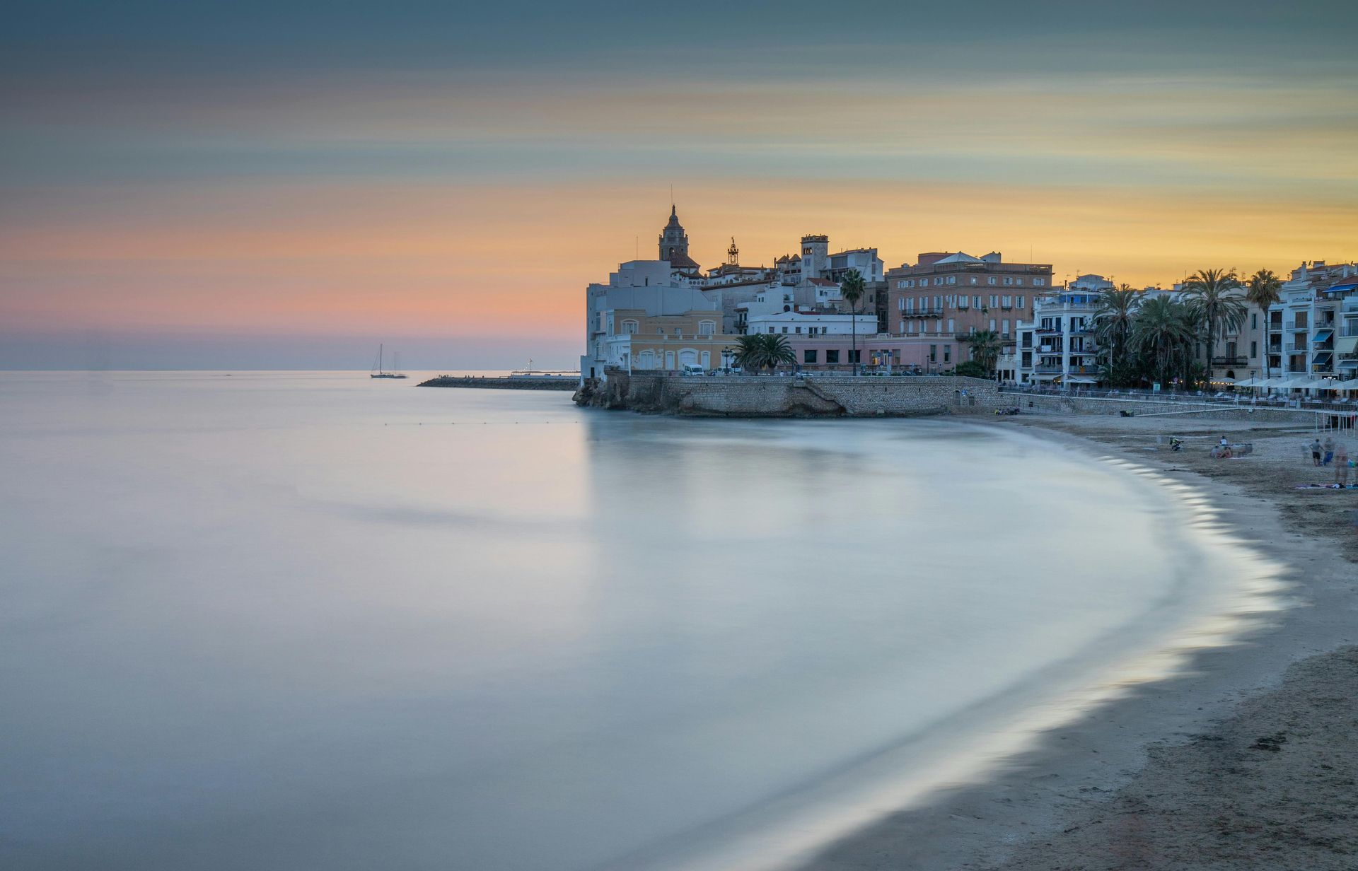 Coastal town at sunset, buildings on a promontory, beach in foreground, calm water, pastel sky.