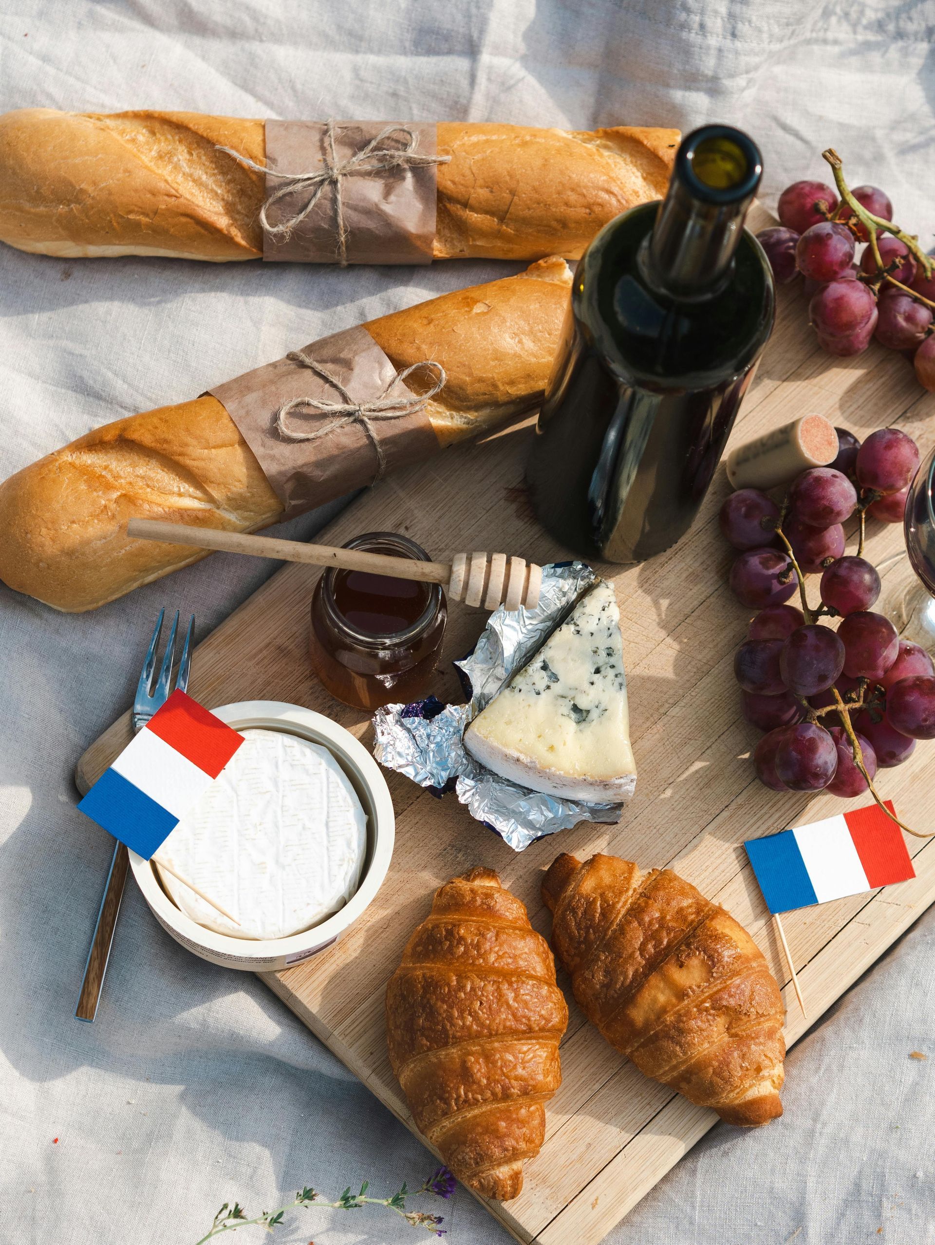 A French picnic spread with baguettes, croissants, blue cheese, grapes, honey, wine, and small French flags on a board.