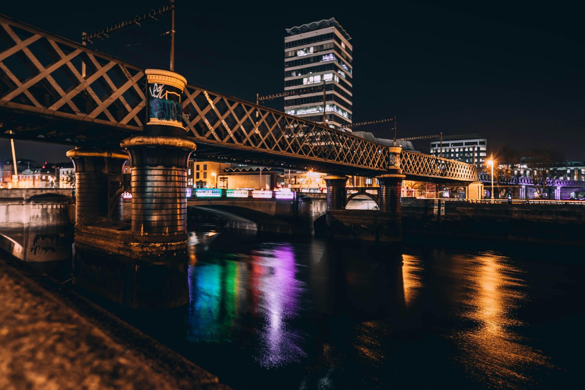 A night view of a dark metal bridge over a river, with colorful light reflections shimmering on the water surface.
