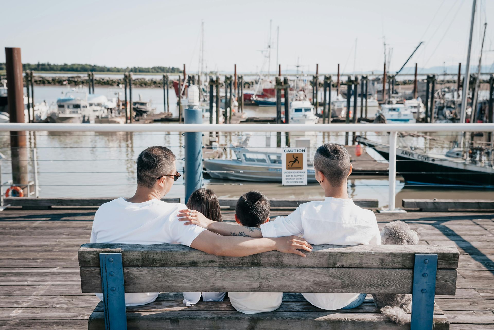 Family of four sits on a bench overlooking a harbor with boats, sunny day.