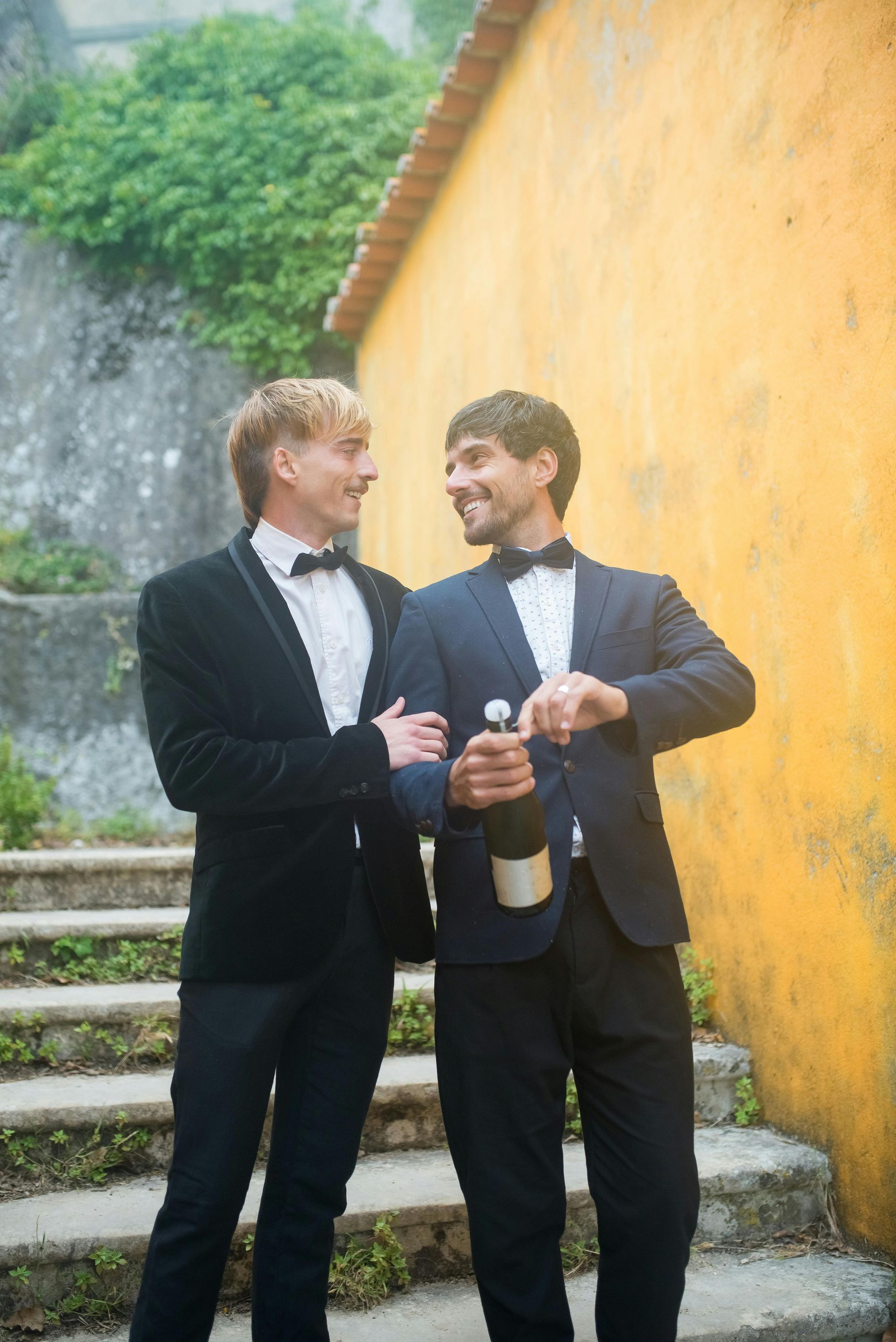 Two men in tuxedos, one opening champagne bottle, smiling at each other by a yellow wall and stone steps.