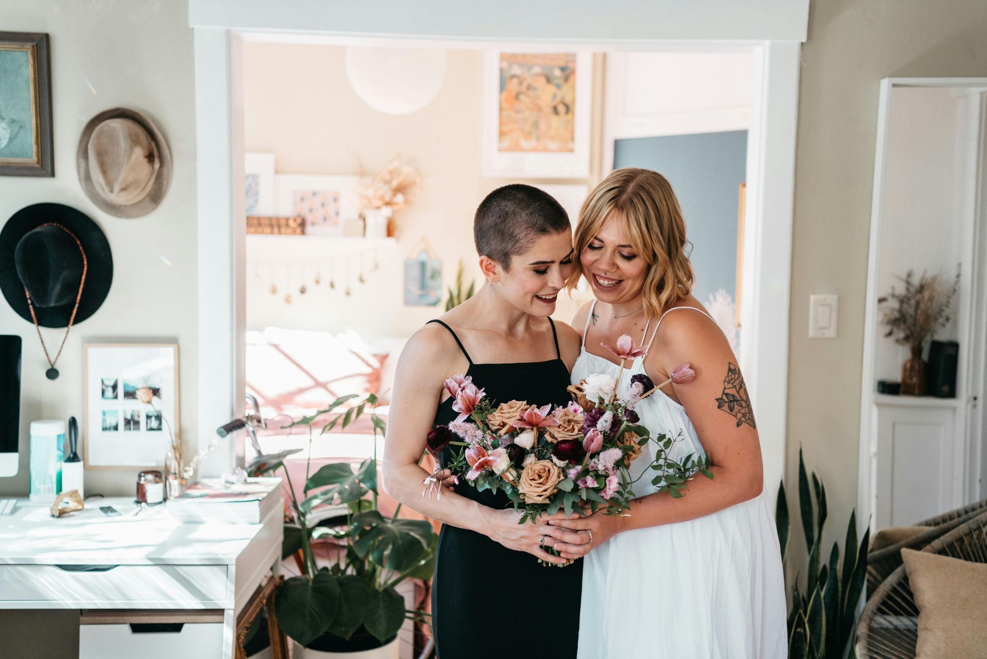Two brides smiling, one with a buzz cut and black dress, the other with long blonde hair in a white dress, holding flowers.