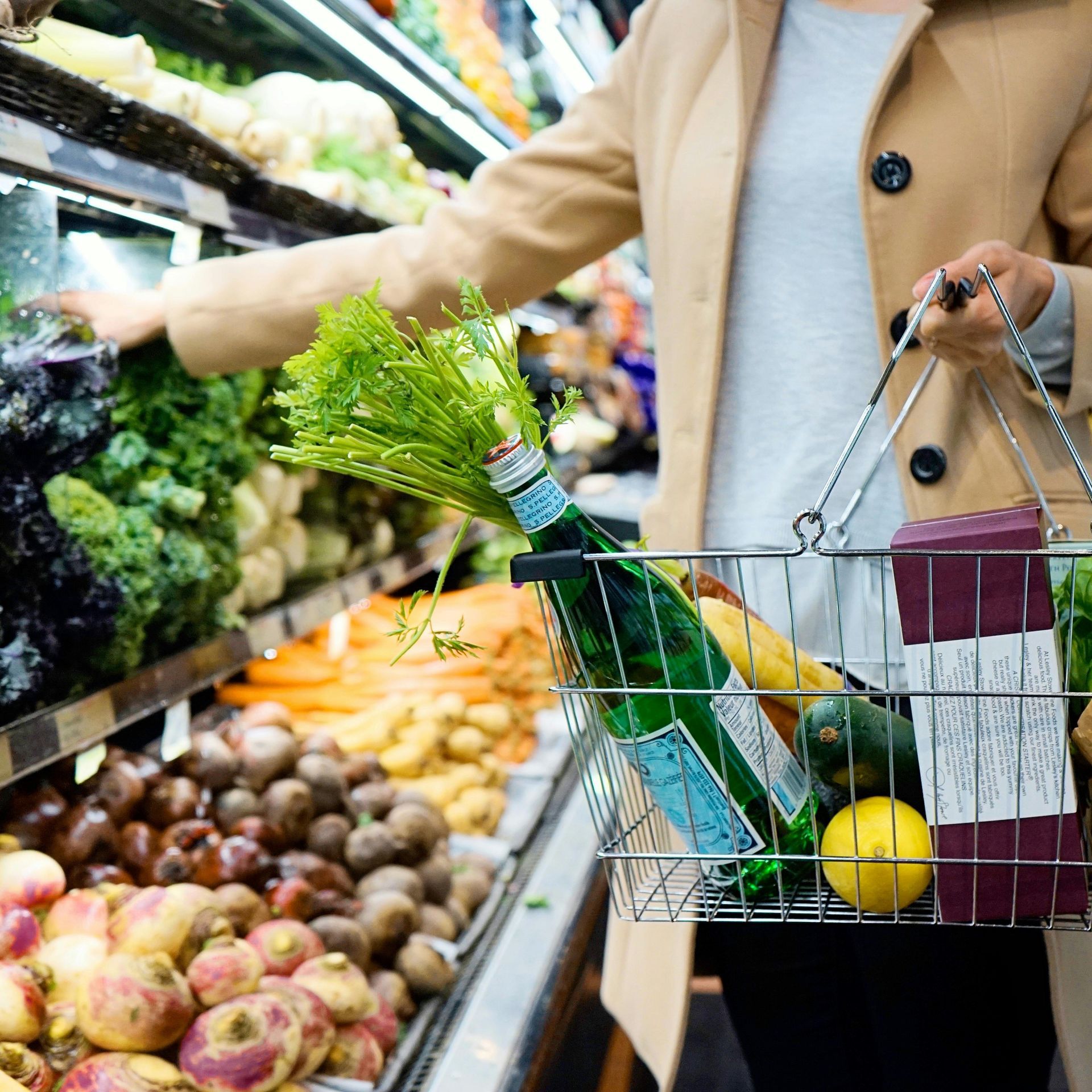 A person holding a wire shopping basket filled with produce and a bottle of sparkling water in a grocery store.