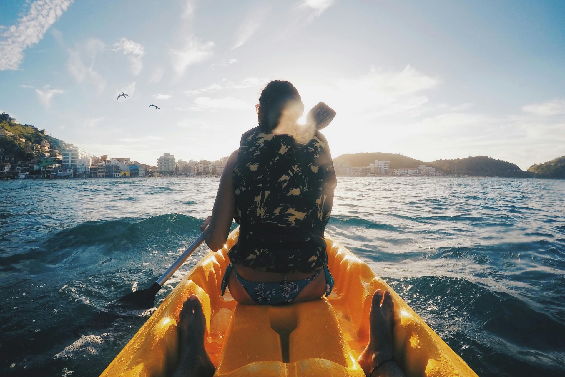 Person kayaking on orange kayak in sunny water, city in background.