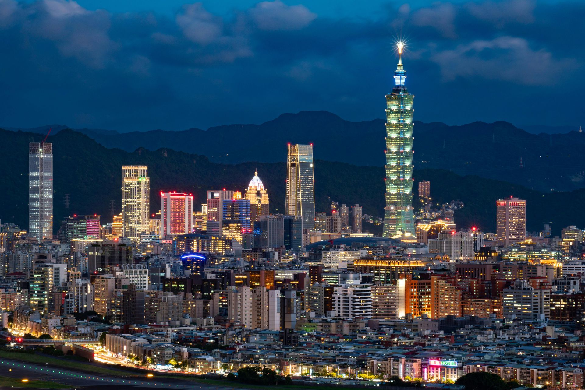 A twilight view of the Taipei skyline, featuring the illuminated Taipei 101 skyscraper against a backdrop of mountains.
