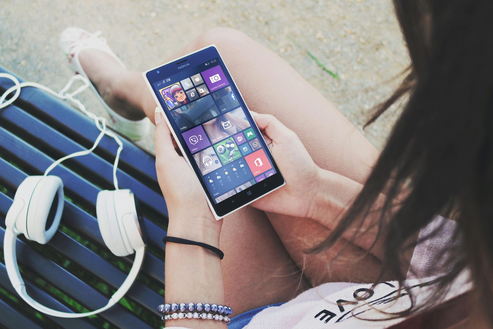 Woman holding a smartphone with the Windows start screen, sitting on a bench with white headphones.