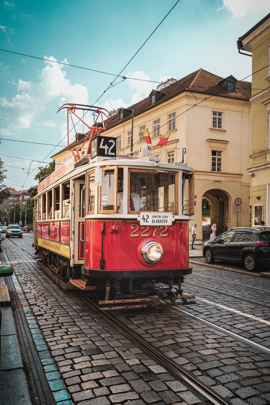 Red tram car on cobblestone street, building in background, overhead wires.