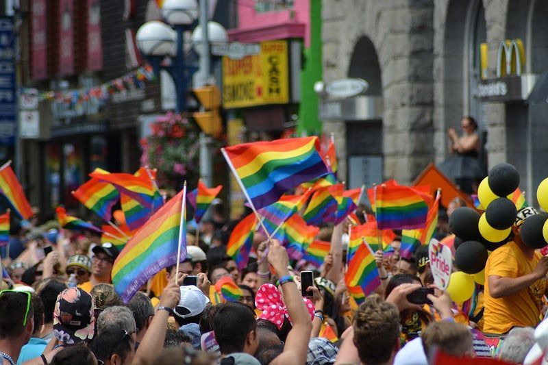 People wave rainbow flags at a Pride parade, celebrating with balloons and street decorations.
