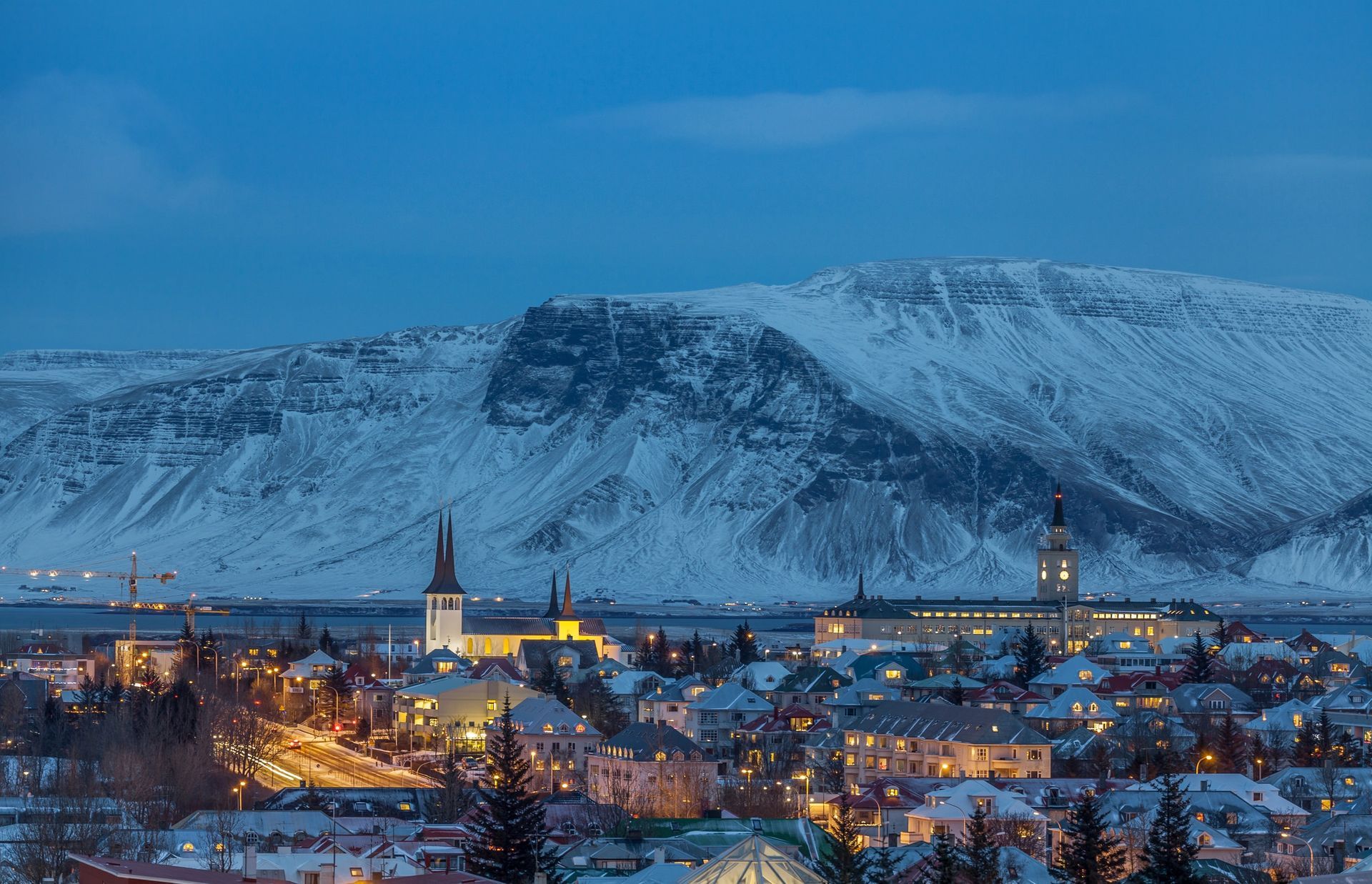 Snowy mountain overlooks Reykjavik, Iceland, with illuminated buildings and a church at dusk.
