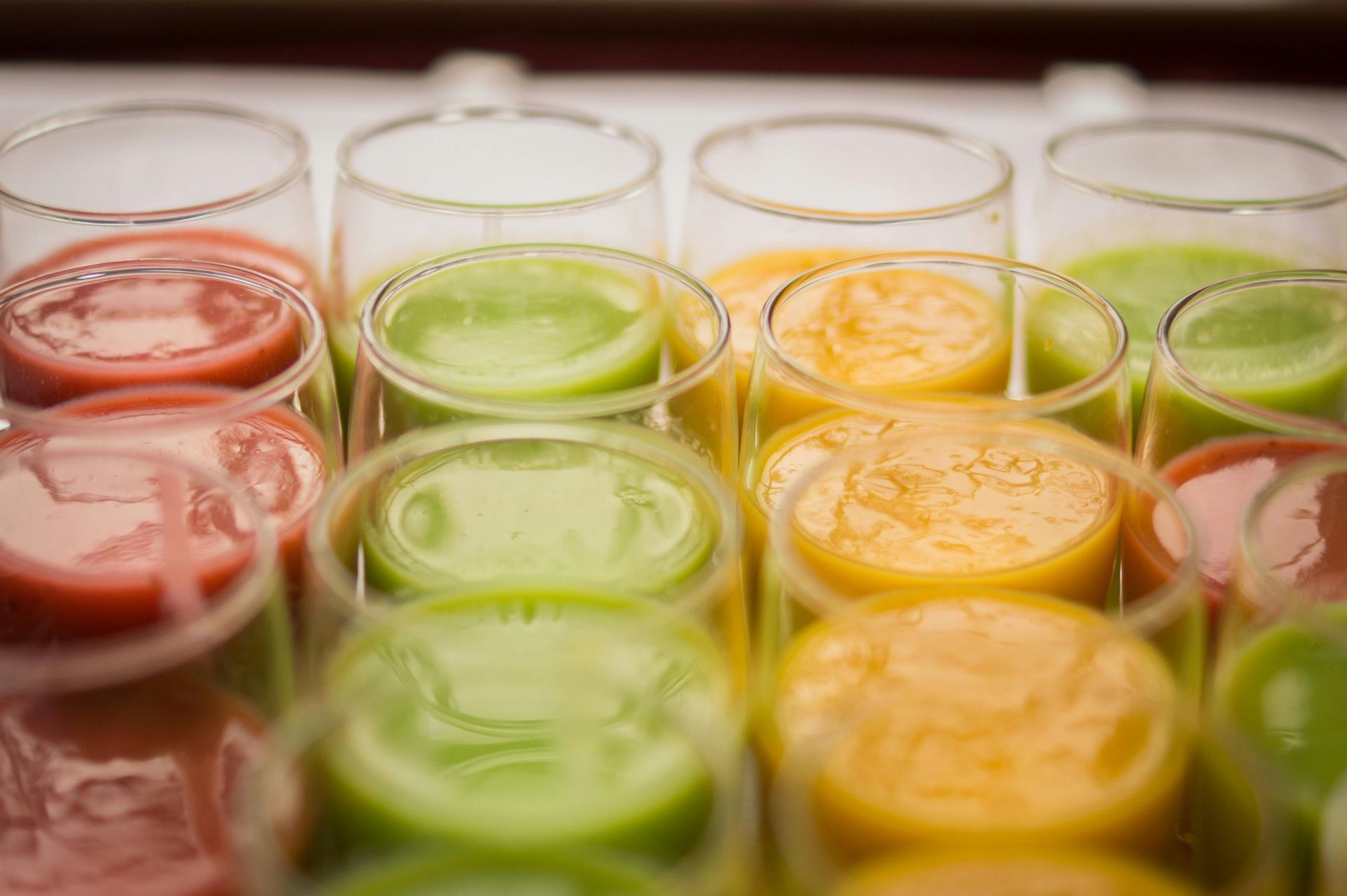 A high-angle view shows rows of glasses filled with red, green, and orange fruit or vegetable smoothies.