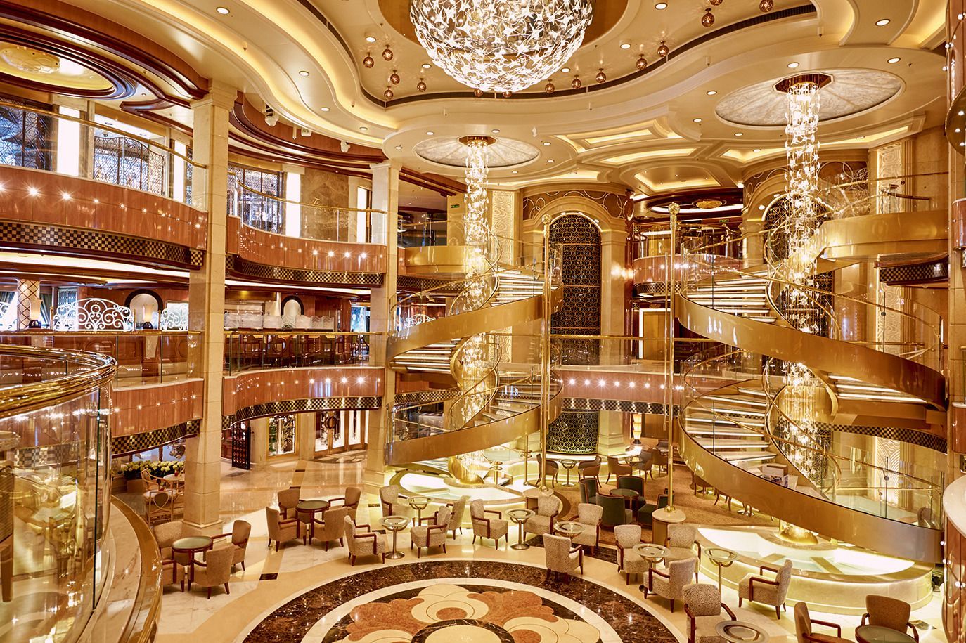 Elegant multi-story cruise ship atrium with gold accents, spiral staircases, and chandeliers.