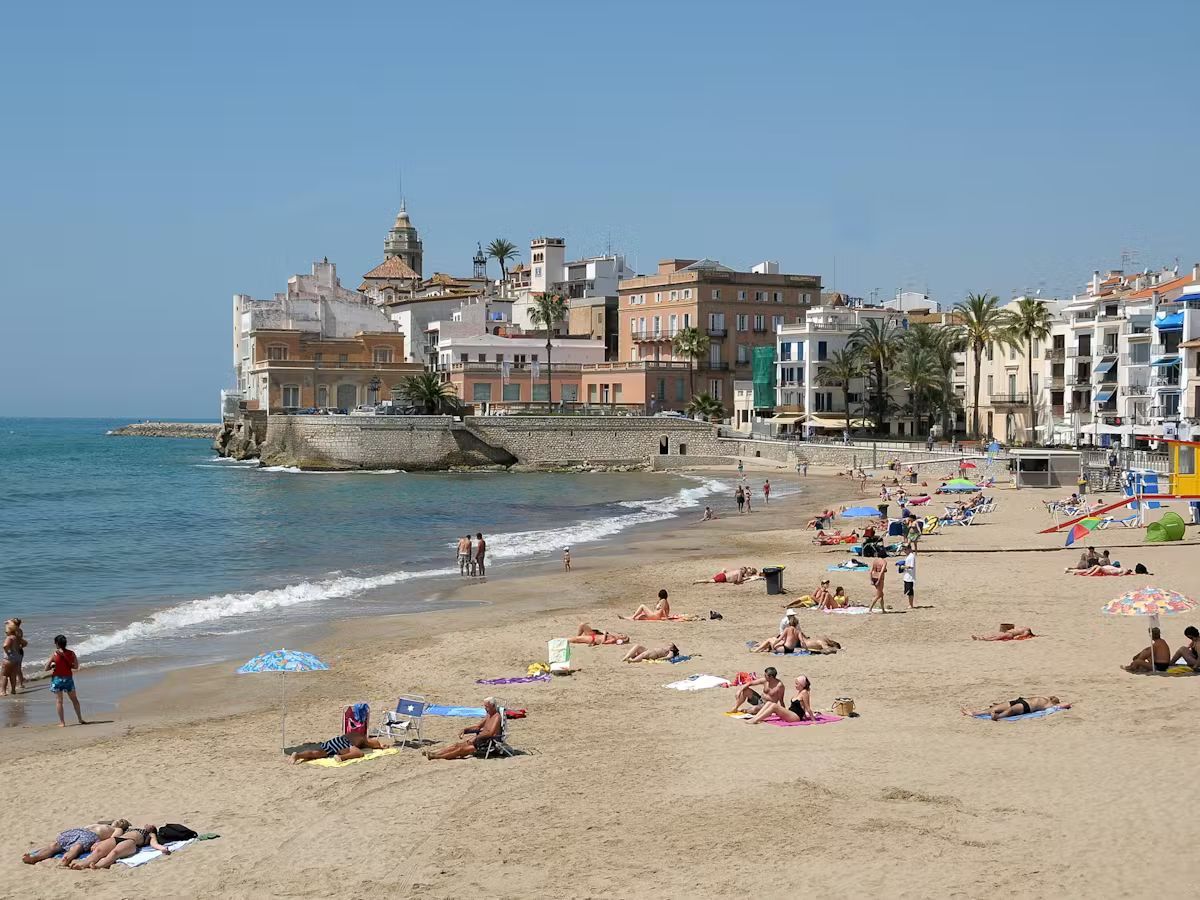 Beach with people sunbathing and swimming; town buildings in the background under a blue sky. Sitges