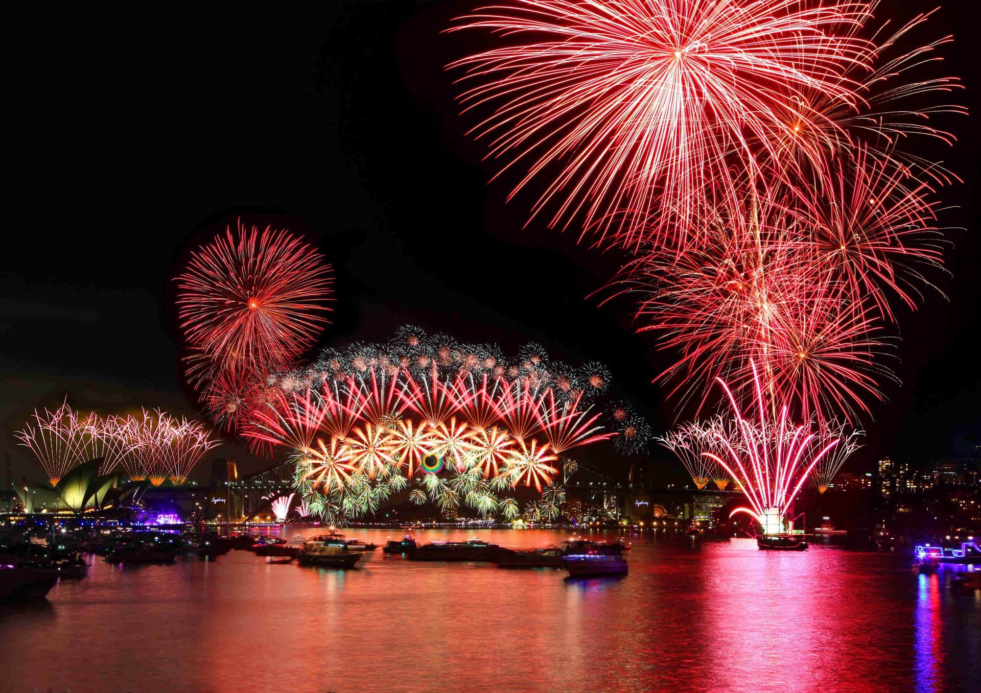 Red fireworks explode over a harbor at night, reflecting in the water.