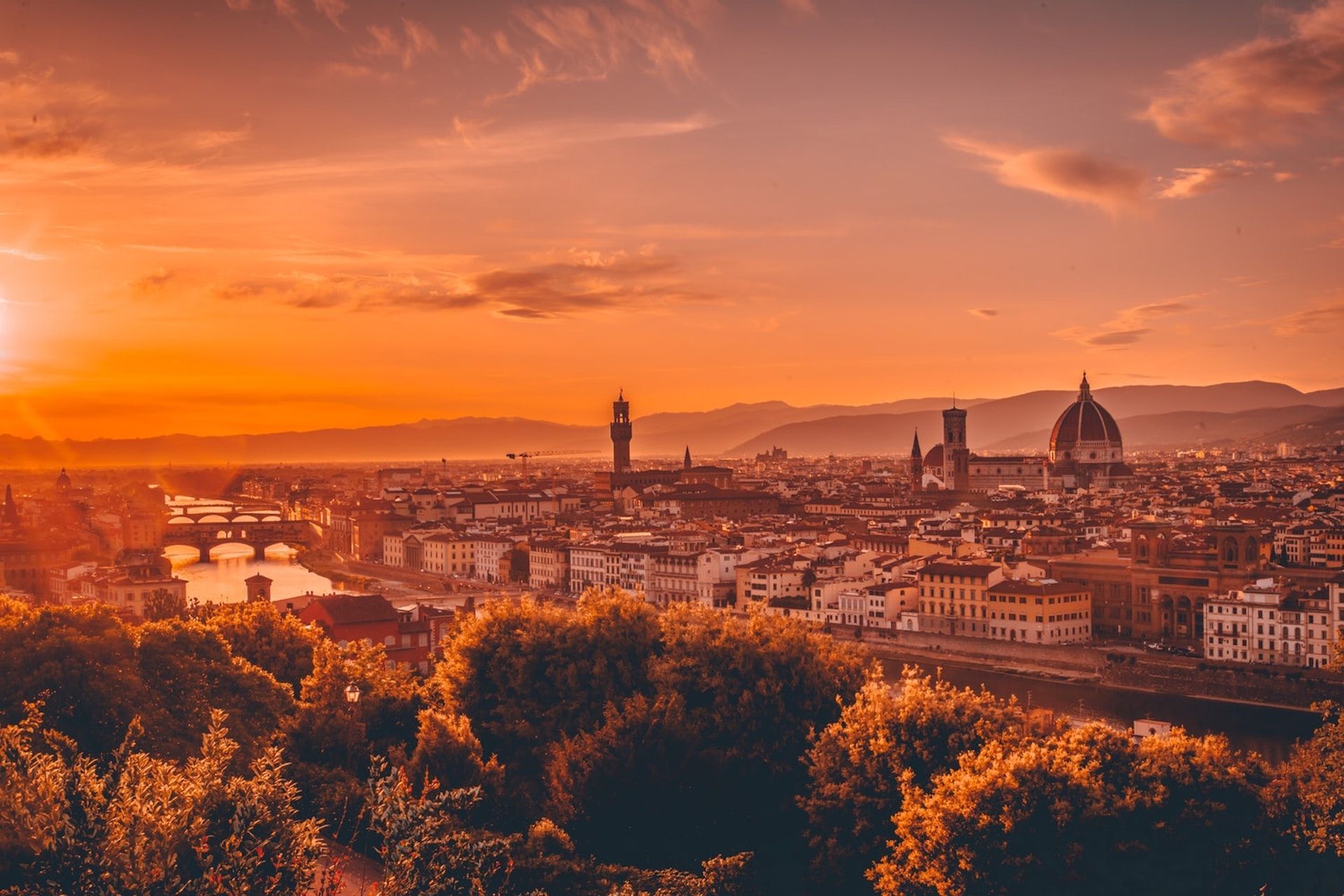 Sunset over Florence, Italy, with the Duomo and Ponte Vecchio silhouetted, bathed in orange and gold light.