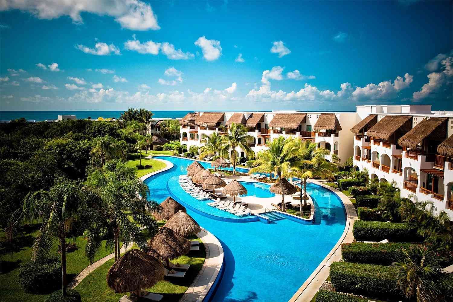 Resort with winding blue pool, thatched roofs, lush green landscaping, and ocean view under a sunny sky.