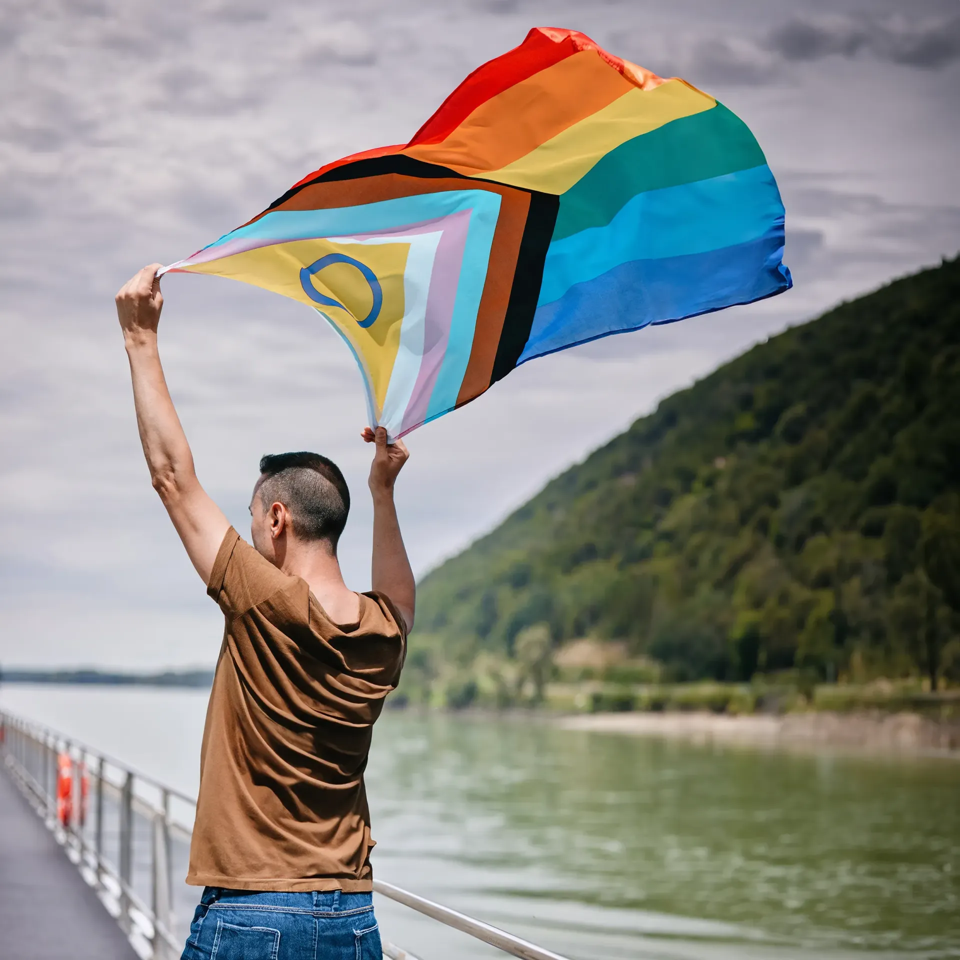 Person waving a rainbow and bisexual pride flag by a riverside walkway