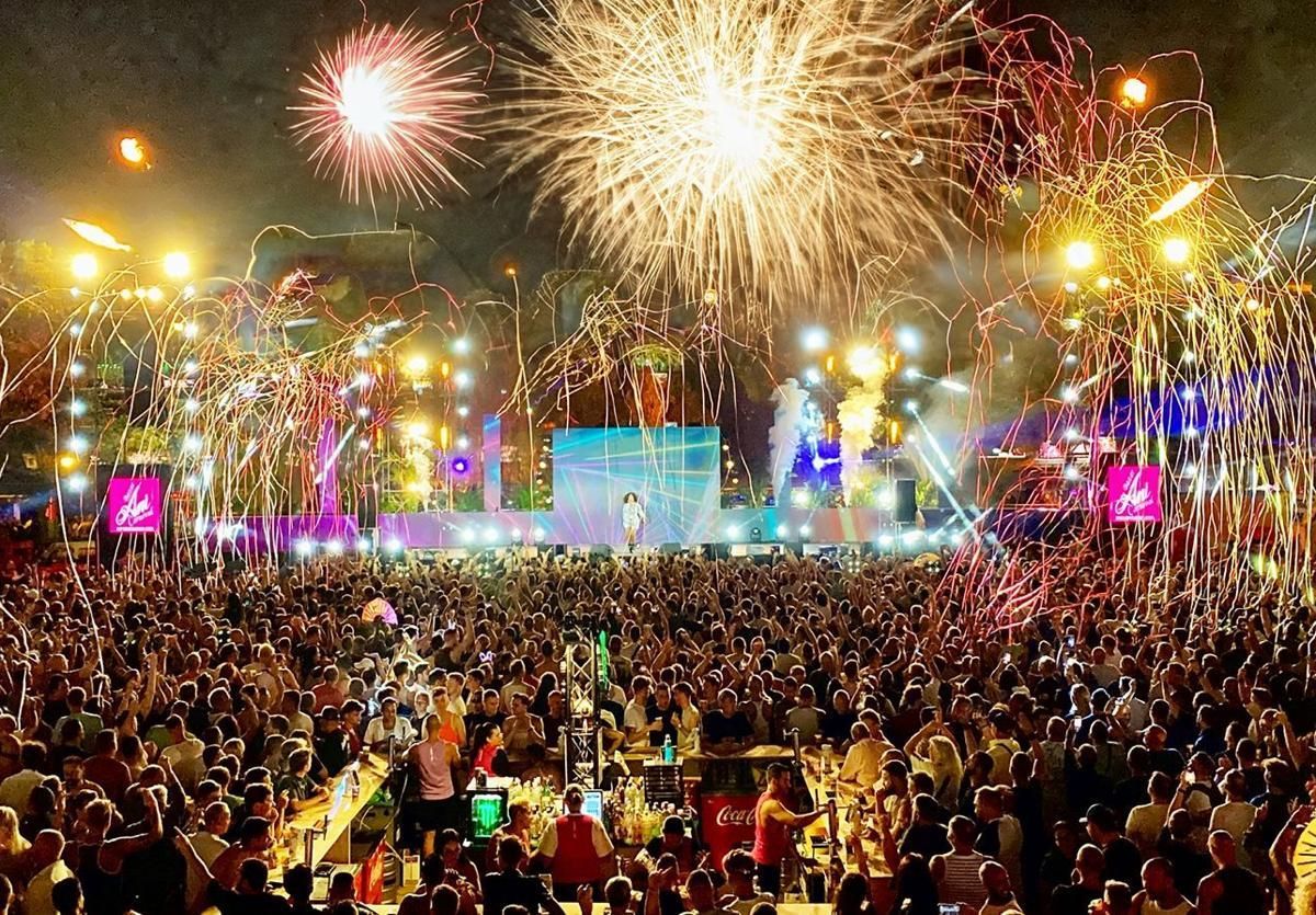 Fireworks explode over a large crowd at a concert stage, lit with bright lights, night.