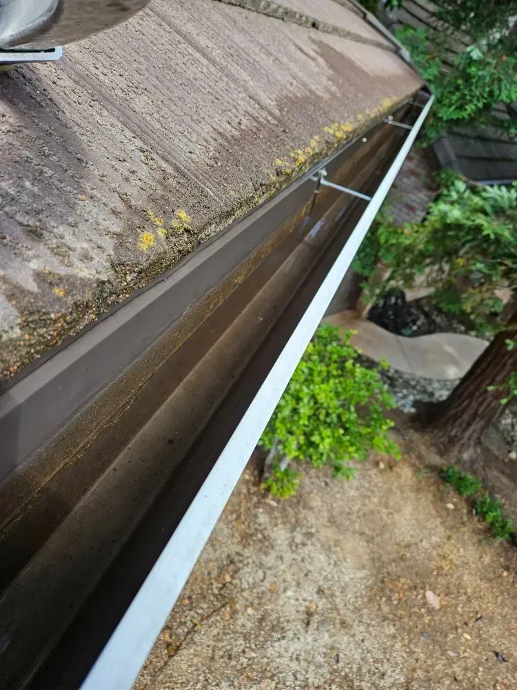 A brown gutter filled with debris, attached to a roof with some vegetation nearby.