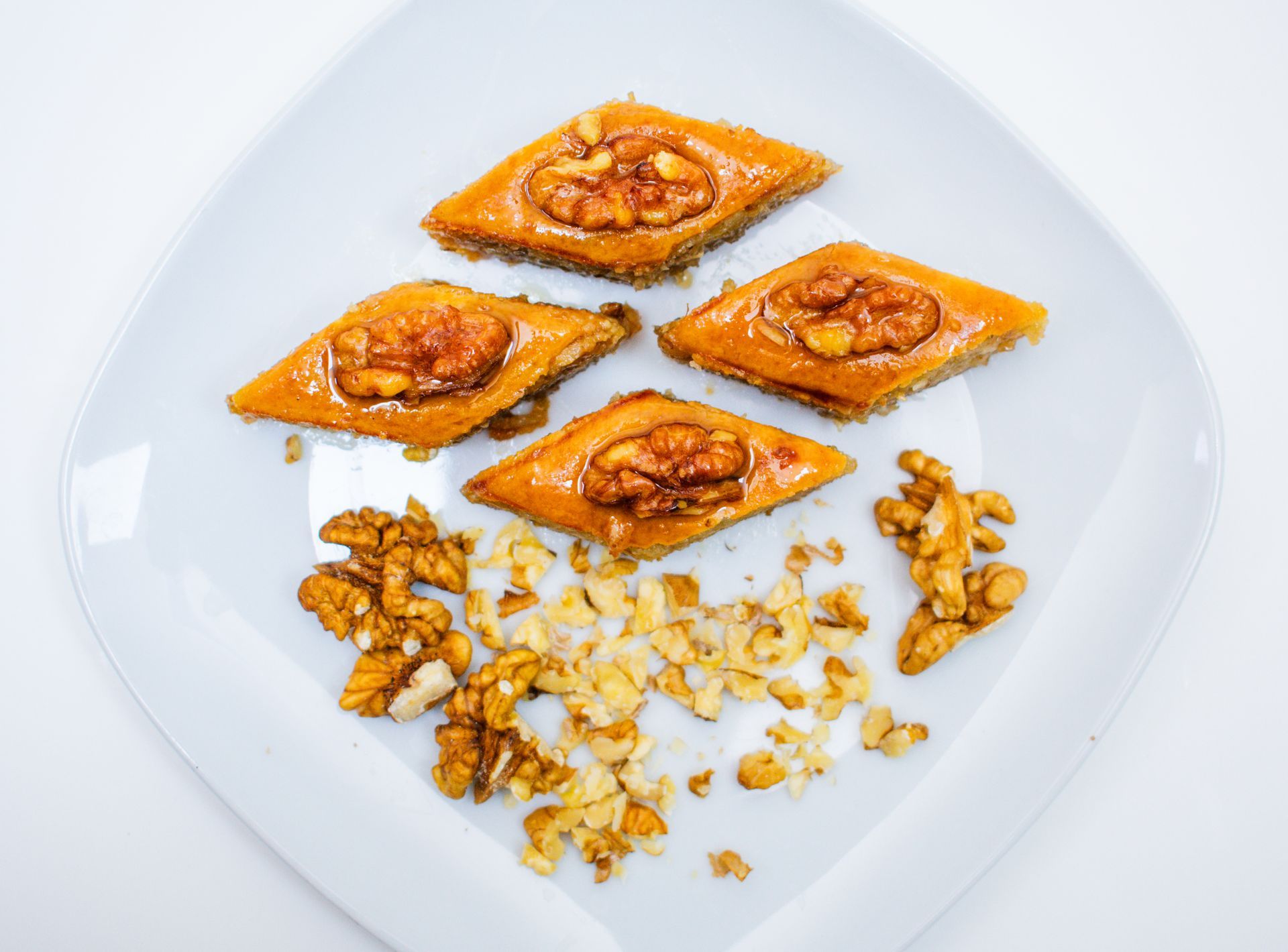 Four diamond-shaped pakhlava pastries topped with walnuts on a white plate, with scattered walnuts.