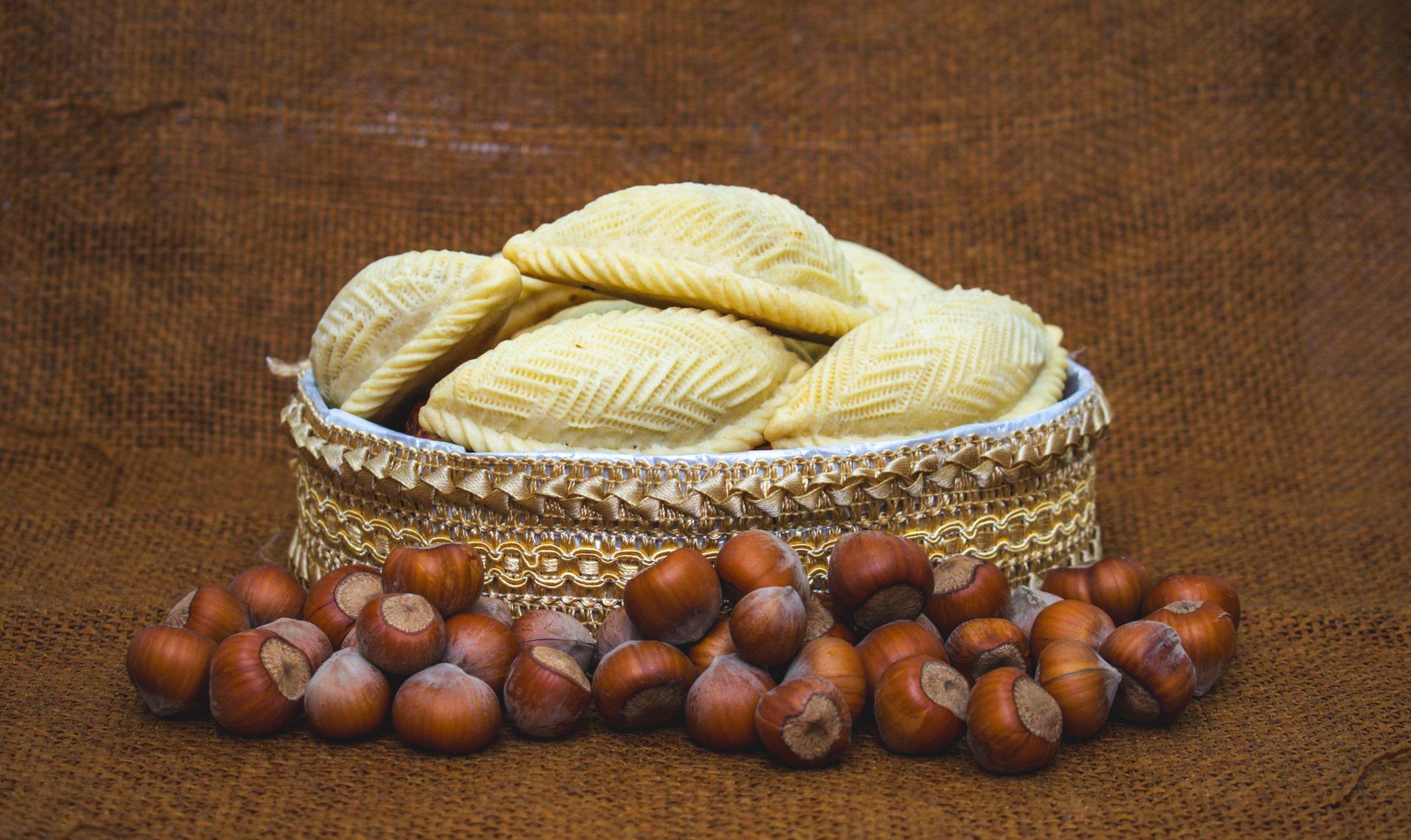 Azerbaijani shekerbura pastries in a woven basket with hazelnuts on a brown cloth.