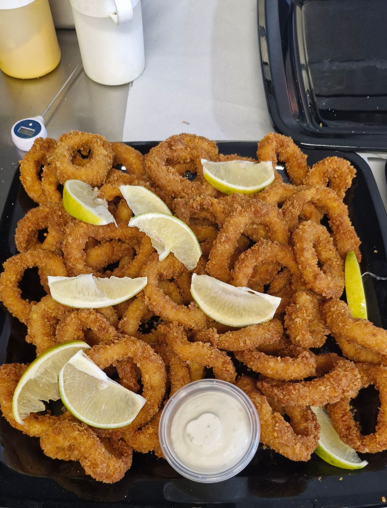 Crispy fried onion rings on a black tray, garnished with lime wedges and a small cup of sauce.