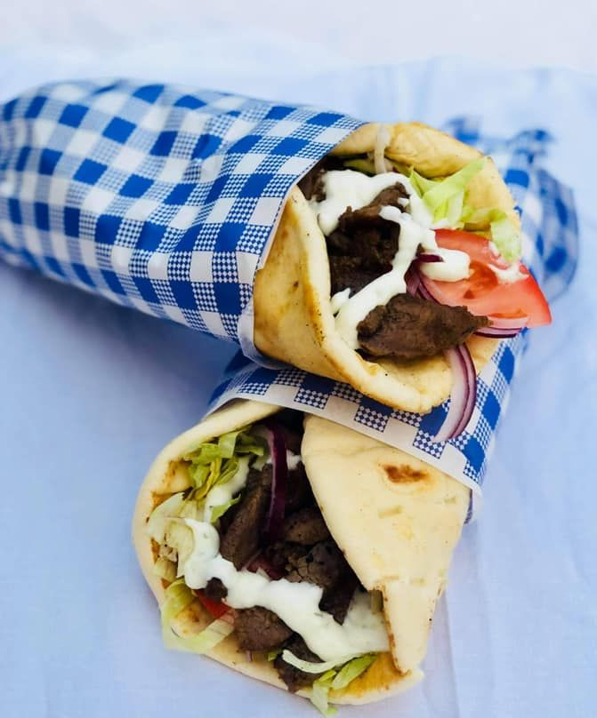 Two gyros wrapped in blue and white checkered paper. Filled with meat, lettuce, tomato, onions, and white sauce.