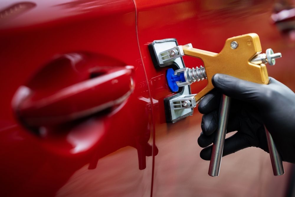 Person using a dent puller tool on a red car door, wearing black gloves.