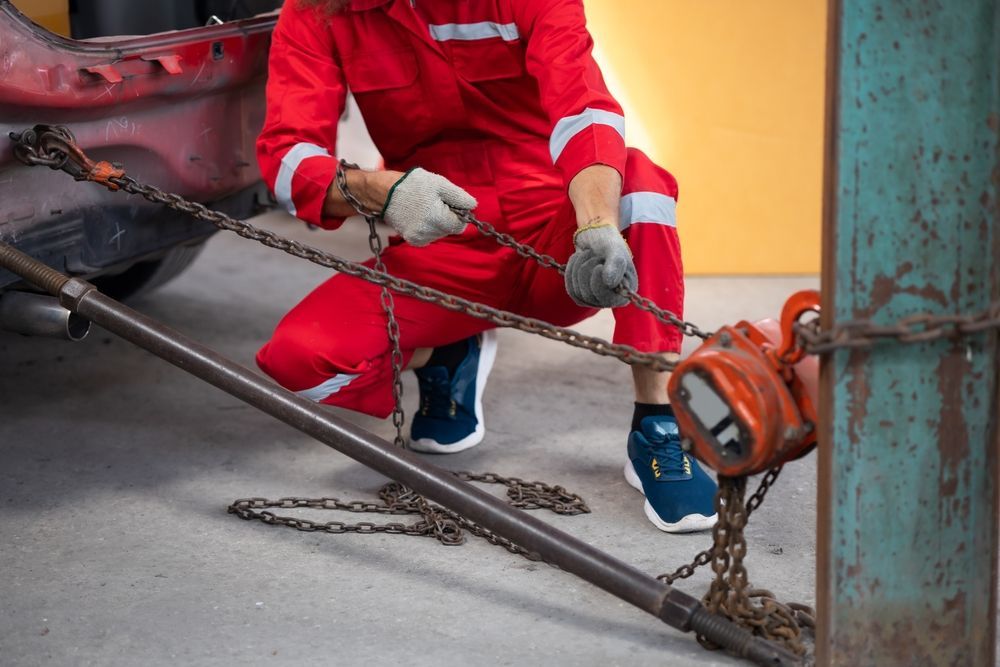 Person in red jumpsuit using a chain hoist to repair a car.