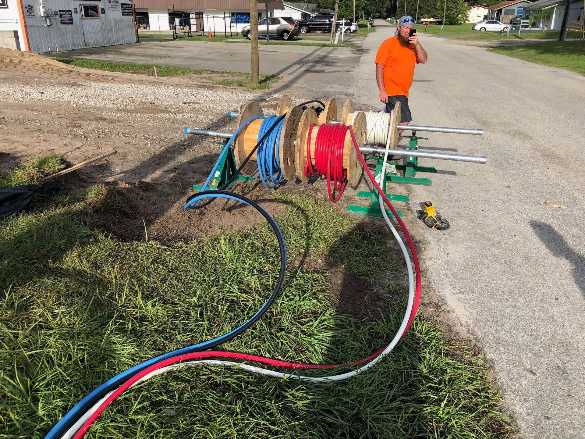 A worker in an orange shirt stands by three wooden spools of colored conduit on a roadside, feeding lines into a trench.