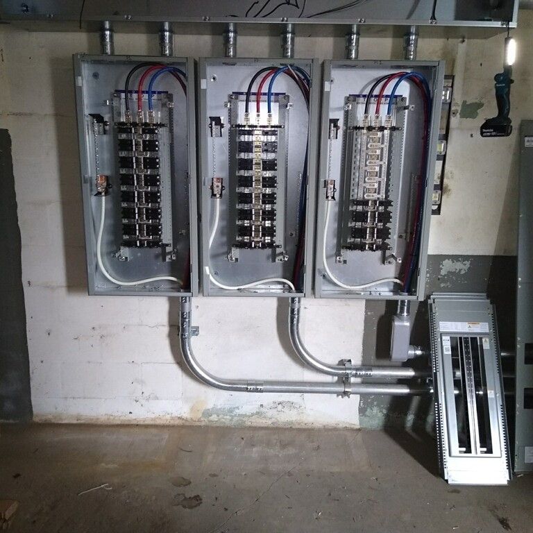 Three electrical panel boxes mounted on a white wall, with wiring connected to the top and conduit pipes along the bottom.