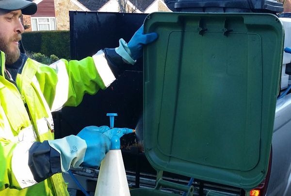 A man is holding a spray bottle in front of a green trash can.