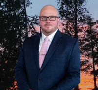Bald man in navy suit and pink tie, wearing glasses, in front of a sunset forest backdrop.