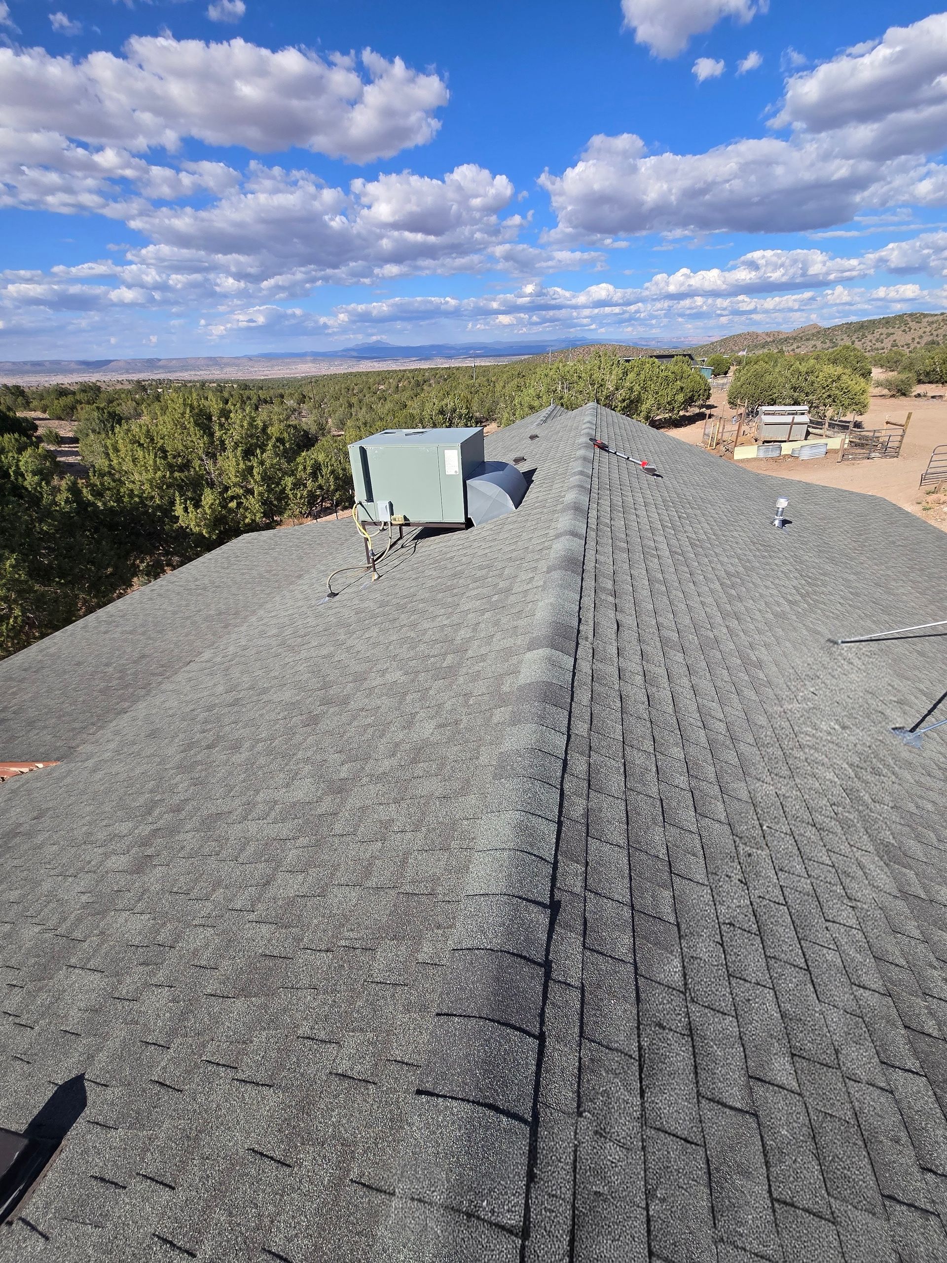 View of a gray asphalt shingle roof with a rooftop unit, trees, and blue sky with clouds.