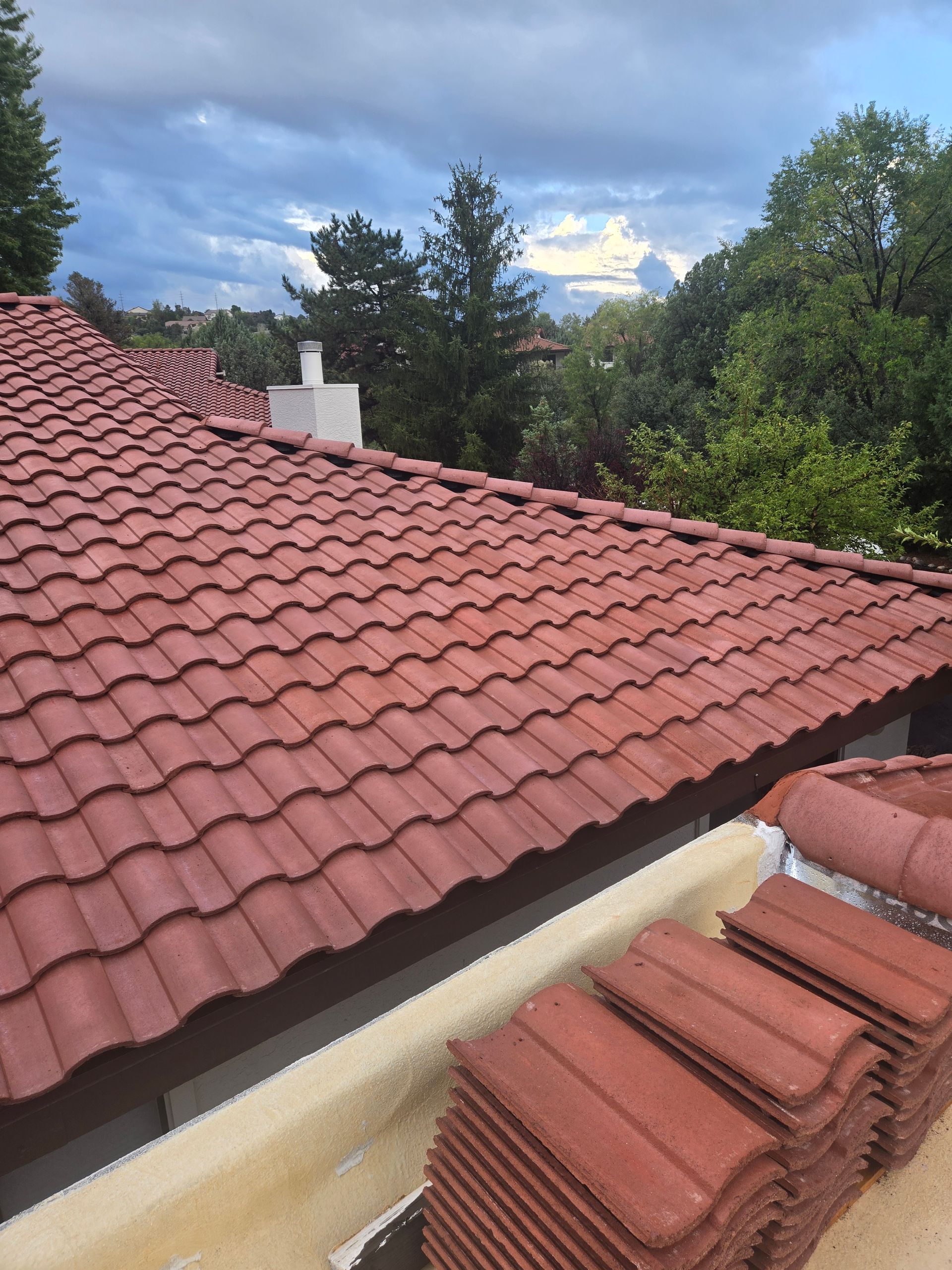Red tile roof with a stack of tiles in foreground; cloudy sky and trees in background.