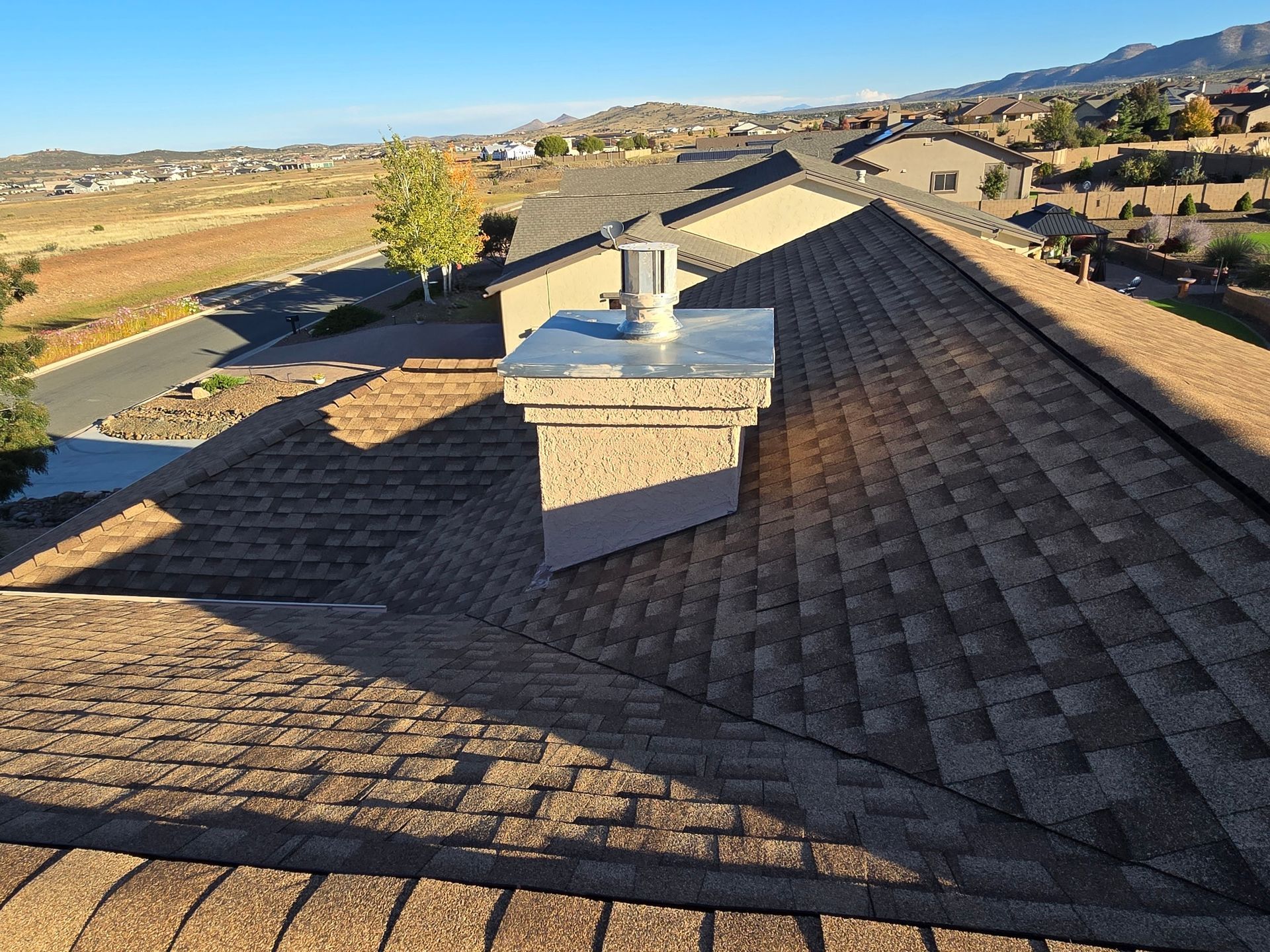 View from rooftop: a chimney with metal cap, brown shingles, houses and landscape in background.